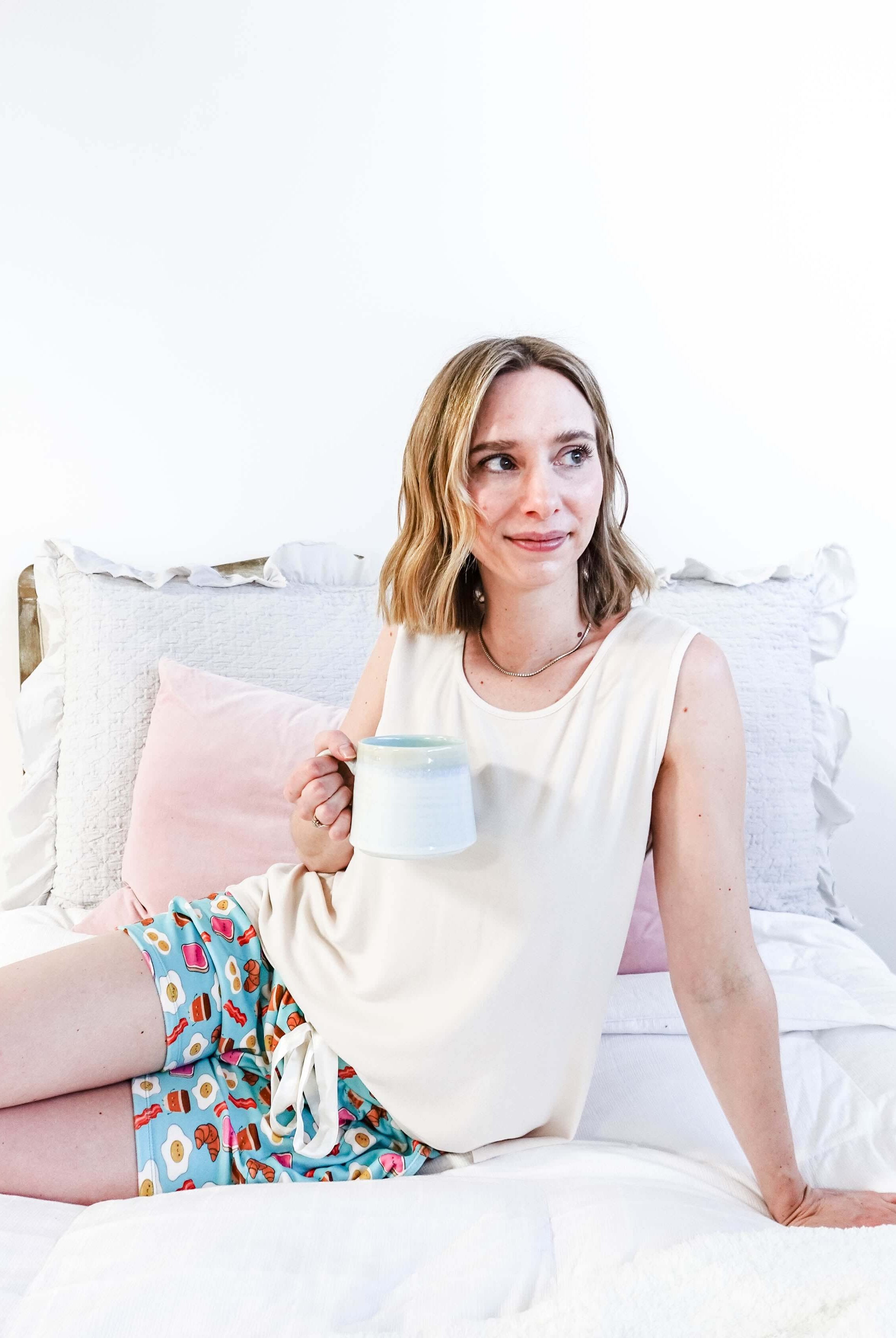 Lifestyle shot of Miranda on a bed wearing the Brunch Club velour shorts with eggs and bacon print and ivory peachy tank.