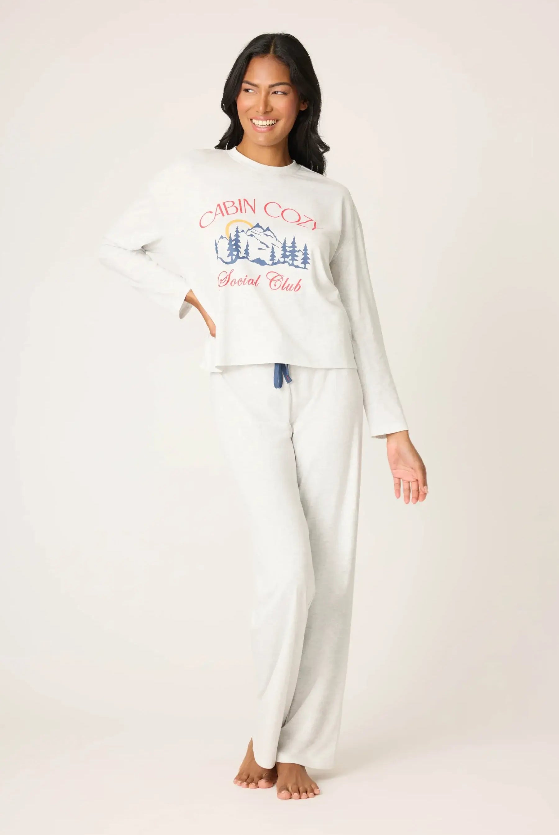 Woman wearing a white long-sleeve top and pants with a cabin cozy social club graphic design on a plain background