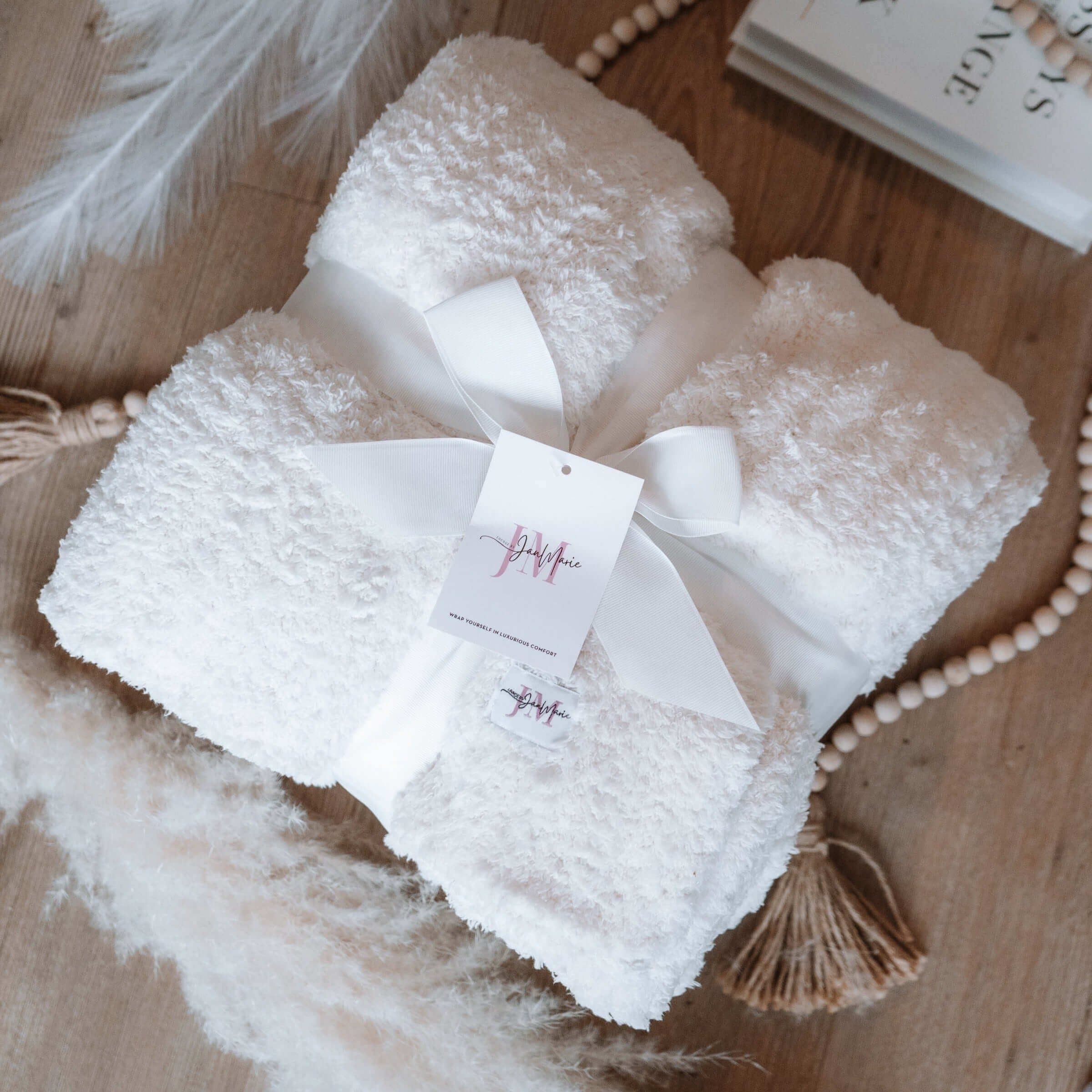 Cozy Cabin Blanket – Oversized Ivory Plush Throw (75″ × 52″). Plush ivory white blanket shown folded in a square and wrapped with a satin ribbon in a lay flat setting. There are wooden beads, books and pampas grass shown around the blanket aesthetically. The label and tag are shown.