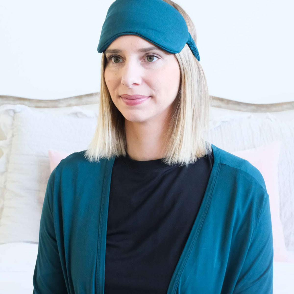A woman with blonde shoulder-length hair wearing a dark teal sleep mask and a matching dark teal cardigan over a black shirt, posed in front of a light-colored bed.