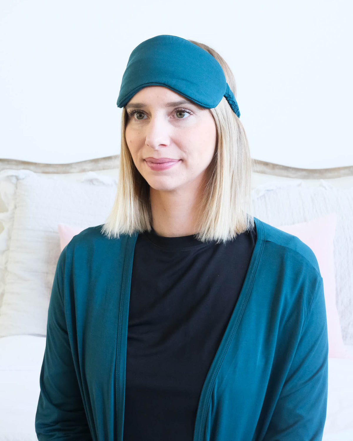 A woman with blonde shoulder-length hair wearing a dark teal sleep mask and a matching dark teal cardigan over a black shirt, posed in front of a light-colored bed.