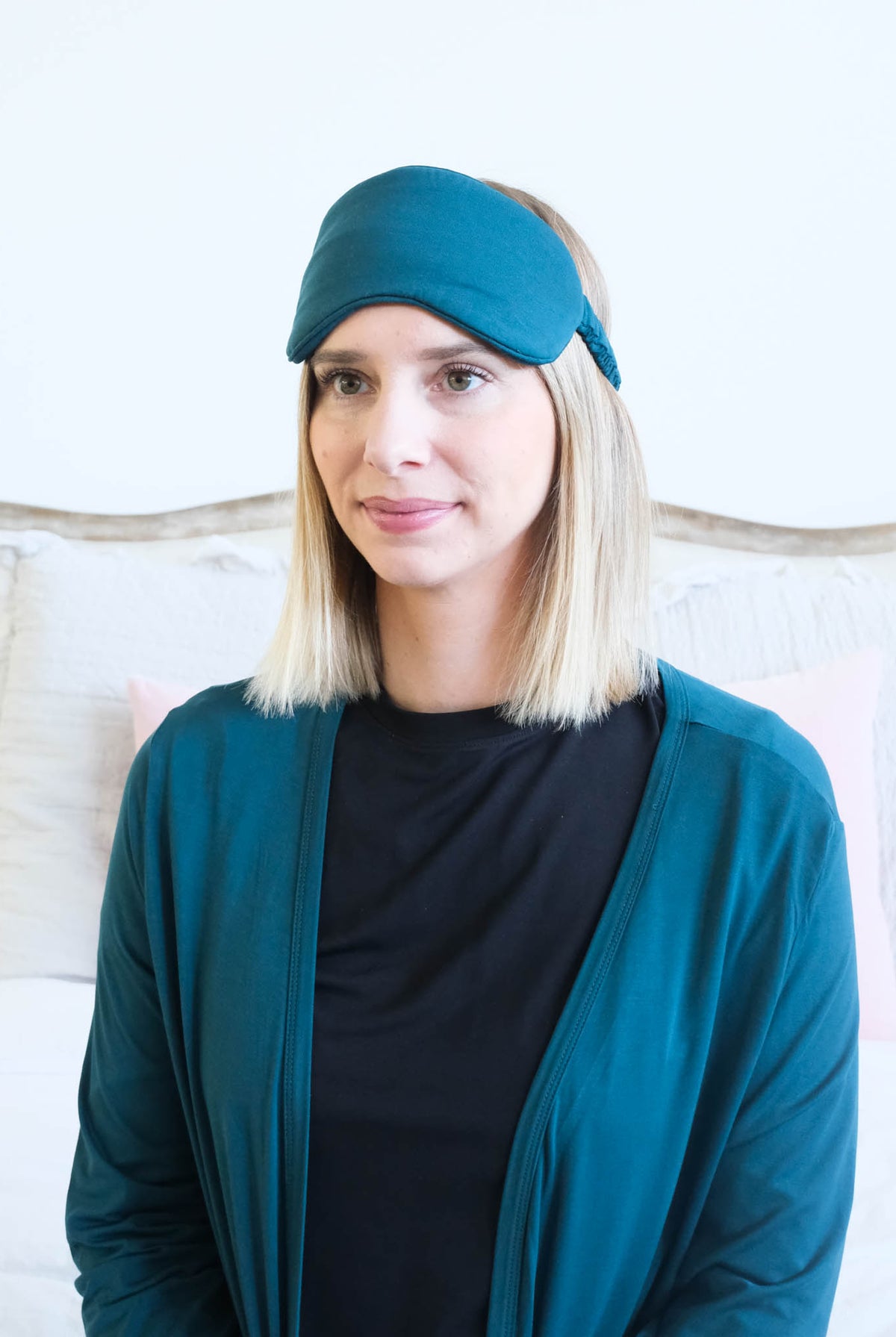A woman with blonde shoulder-length hair wearing a dark teal sleep mask and a matching dark teal cardigan over a black shirt, posed in front of a light-colored bed.
