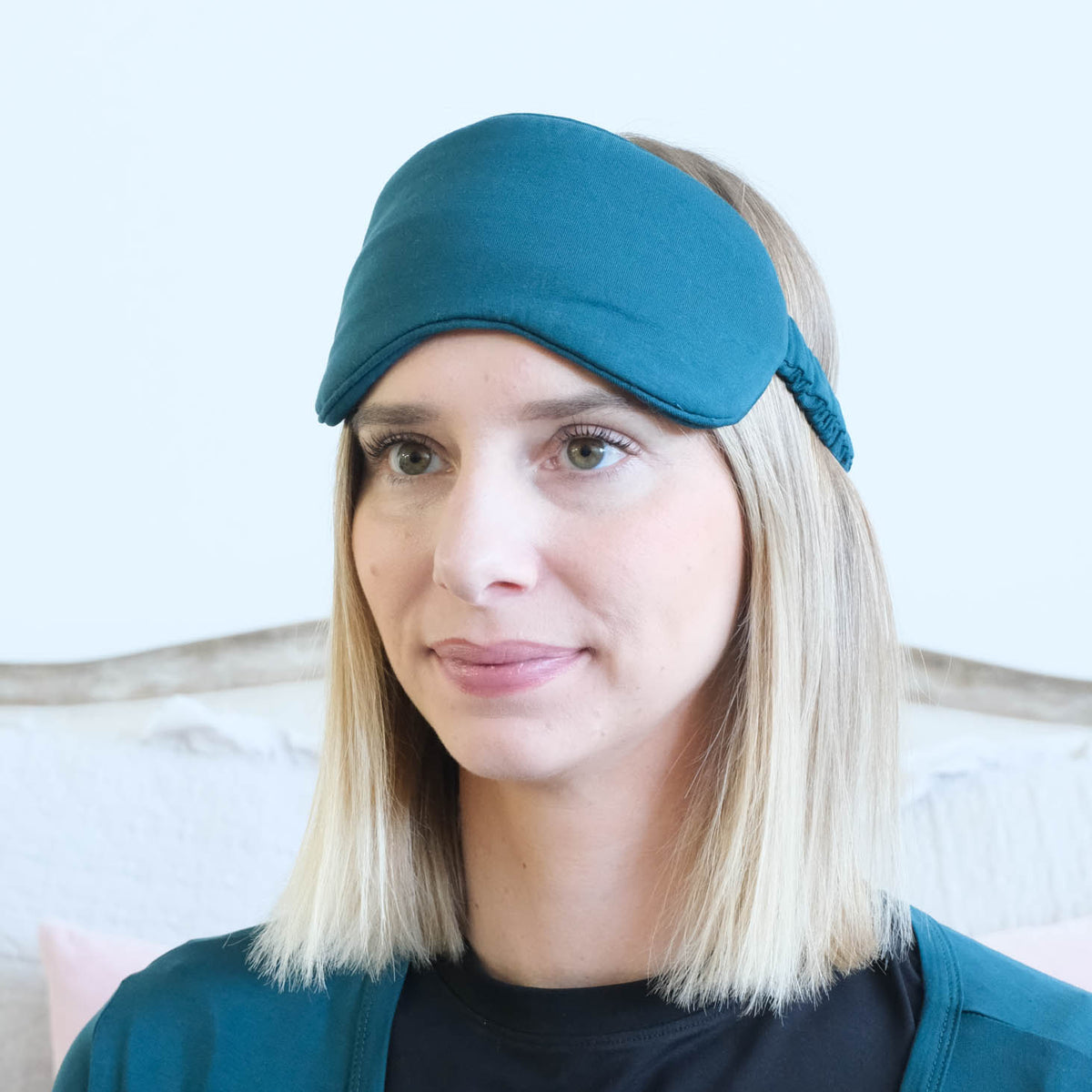 A woman with blonde hair wearing a dark teal sleep mask and a matching dark teal cardigan over a black shirt, set against a light-colored wall and a wooden headboard.