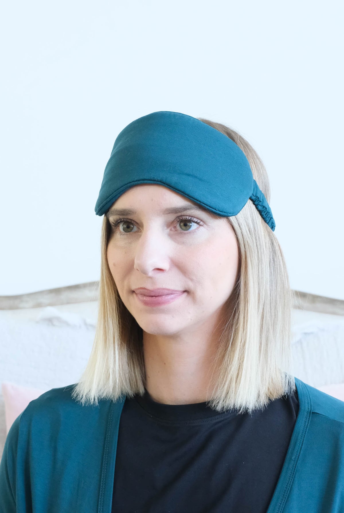 A woman with blonde hair wearing a dark teal sleep mask and a matching dark teal cardigan over a black shirt, set against a light-colored wall and a wooden headboard.