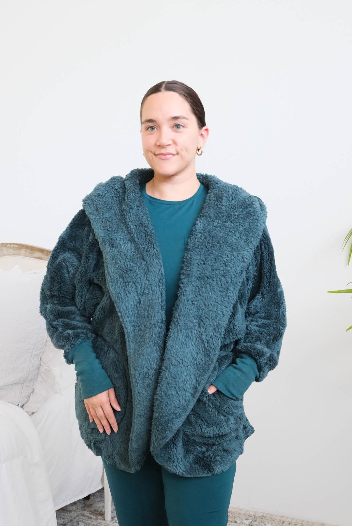 Close up of woman standing in a bedroom wearing a dark peacock teal fluffy hooded cardigan by Lounge by Jan Marie over a dark midnight teal bamboo pajamas, with ribbed cuffs and pink fuzzy slippers, hands tucked into the cardigan's front pockets. Cloud Nine Pajamas Edmonton