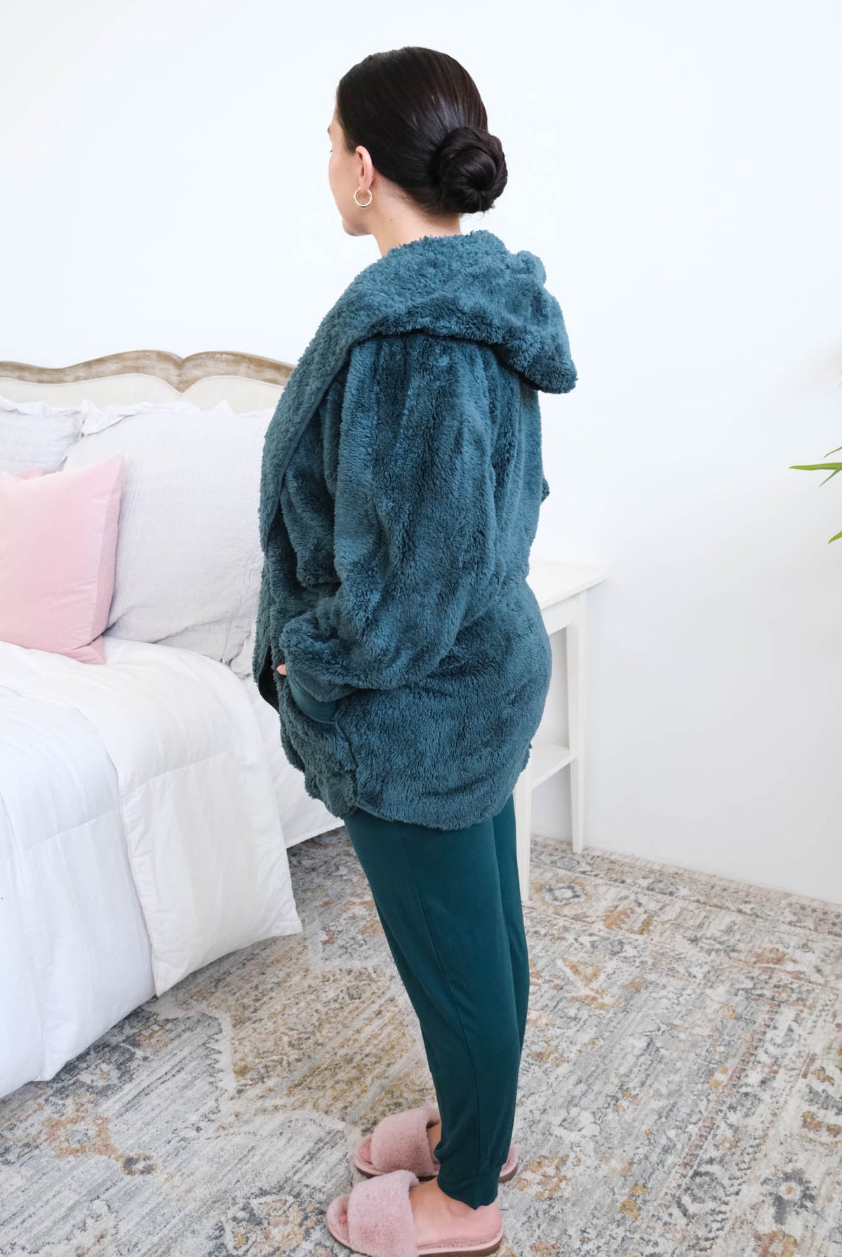 Women standing sideways in a bedroom wearing a dark peacock teal fluffy hooded cardigan by Lounge by Jan Marie over a dark midnight teal bamboo pajamas, with ribbed cuffs and pink fuzzy slippers, hands tucked into the cardigan's front pockets. Cloud Nine Pajamas Edmonton