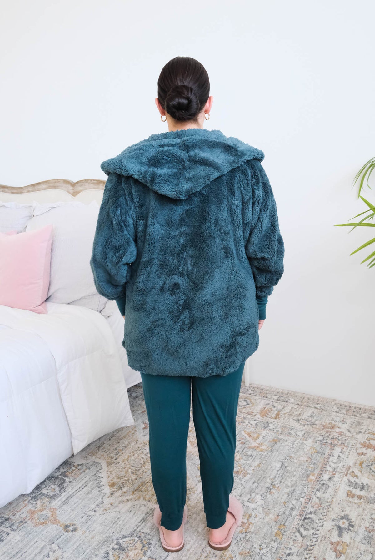 Woman standing backwards in a bedroom wearing a dark peacock teal fluffy hooded cardigan by Lounge by Jan Marie over a dark midnight teal bamboo pajamas, with ribbed cuffs and pink fuzzy slippers, hands tucked into the cardigan's front pockets. Cloud Nine Pajamas Edmonton