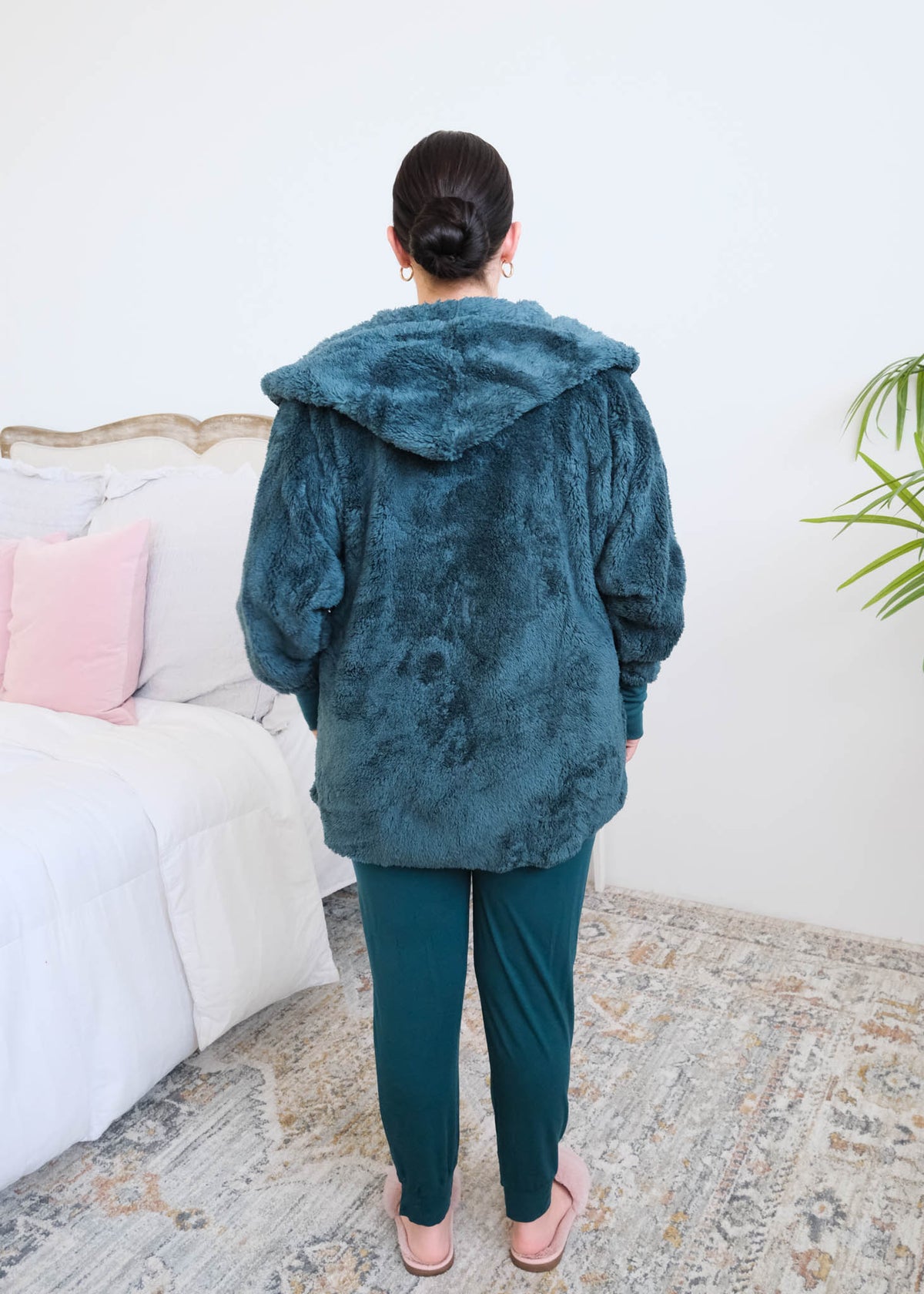 Woman standing backwards in a bedroom wearing a dark peacock teal fluffy hooded cardigan by Lounge by Jan Marie over a dark midnight teal bamboo pajamas, with ribbed cuffs and pink fuzzy slippers, hands tucked into the cardigan's front pockets. Cloud Nine Pajamas Edmonton