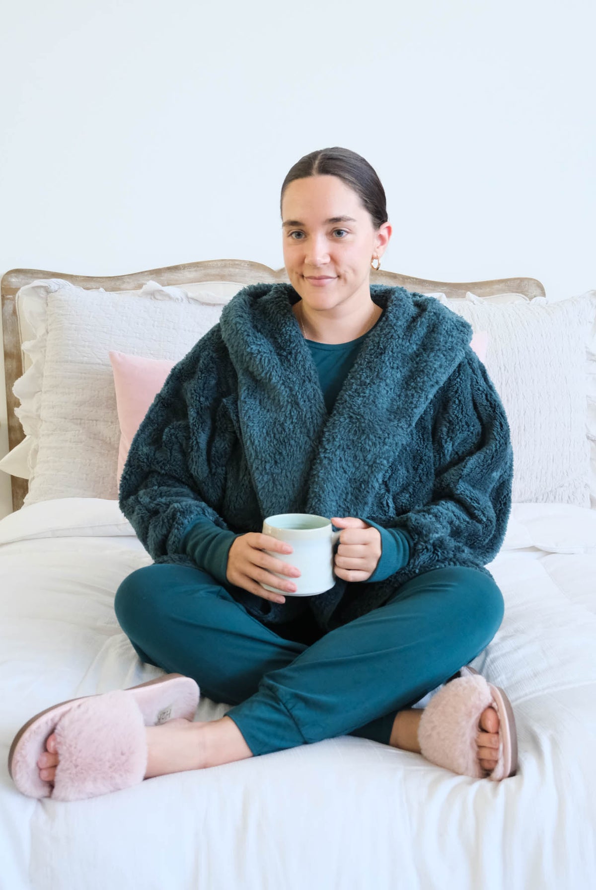 A woman sits cross-legged on a white bed wearing a cozy dark teal fuzzy hooded and matching bamboo teal pajama set, holding a white mug. She has pink slippers on her feet, with white and light pink pillows behind her. Cloud Nine Pajamas Edmonton