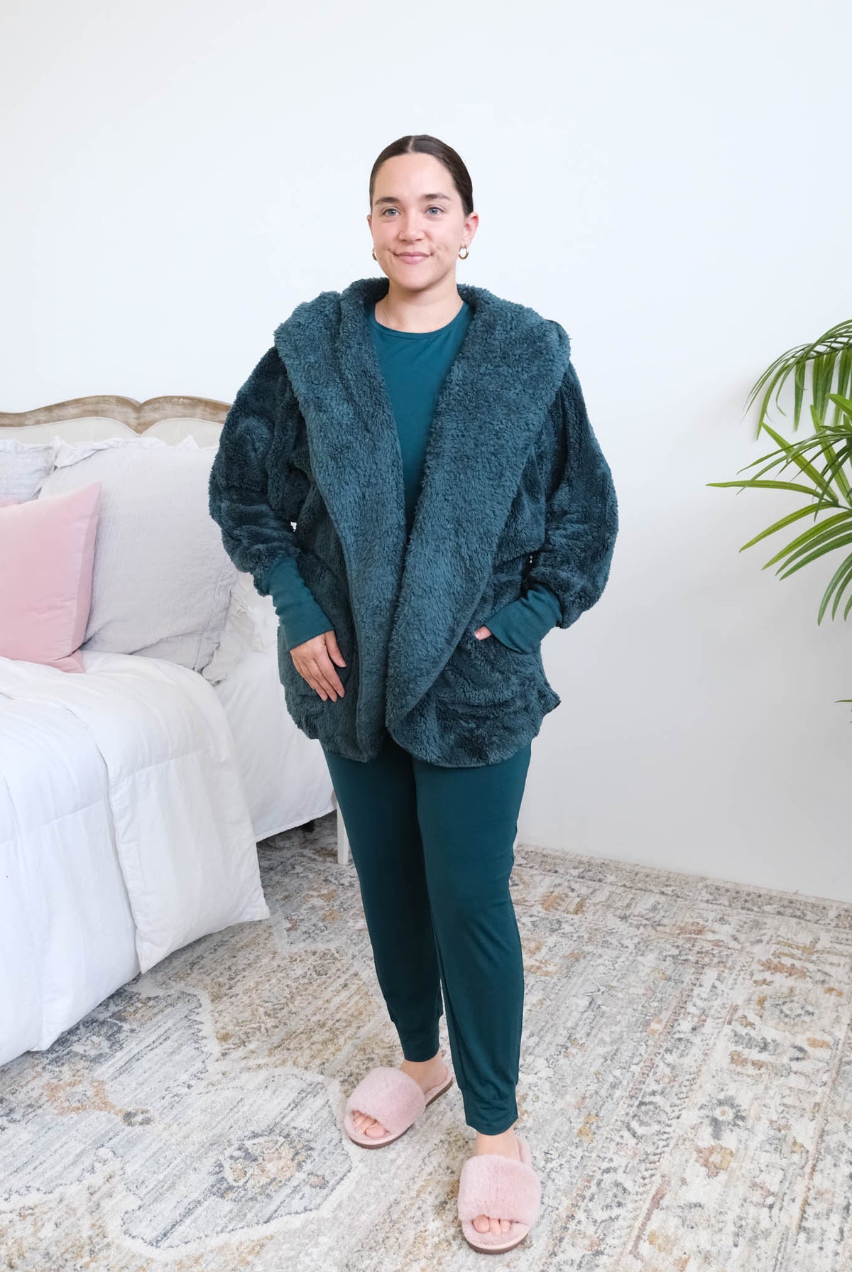 Woman standing in a bedroom wearing a dark peacock teal fluffy hooded cardigan by Lounge by Jan Marie over a dark midnight teal bamboo pajamas, with ribbed cuffs and pink fuzzy slippers, hands tucked into the cardigan's front pockets. Cloud Nine Pajamas Edmonton