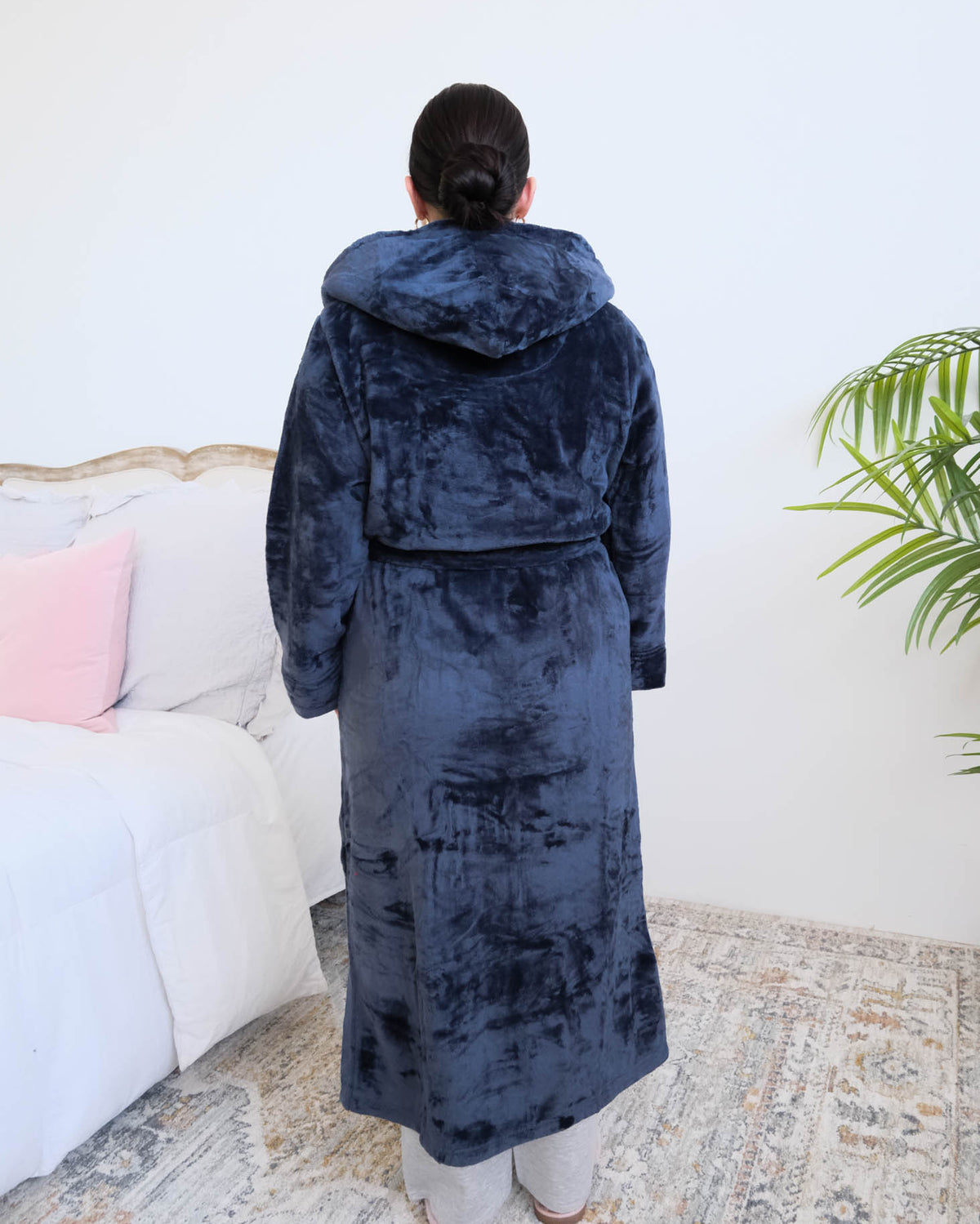 A woman wearing a plush navy blue hooded robe by Lounge By Jan Marie stands backwards in a bedroom setting with a bed, rug, and plant visible in the background. Cloud Nine Pajamas Edmonton