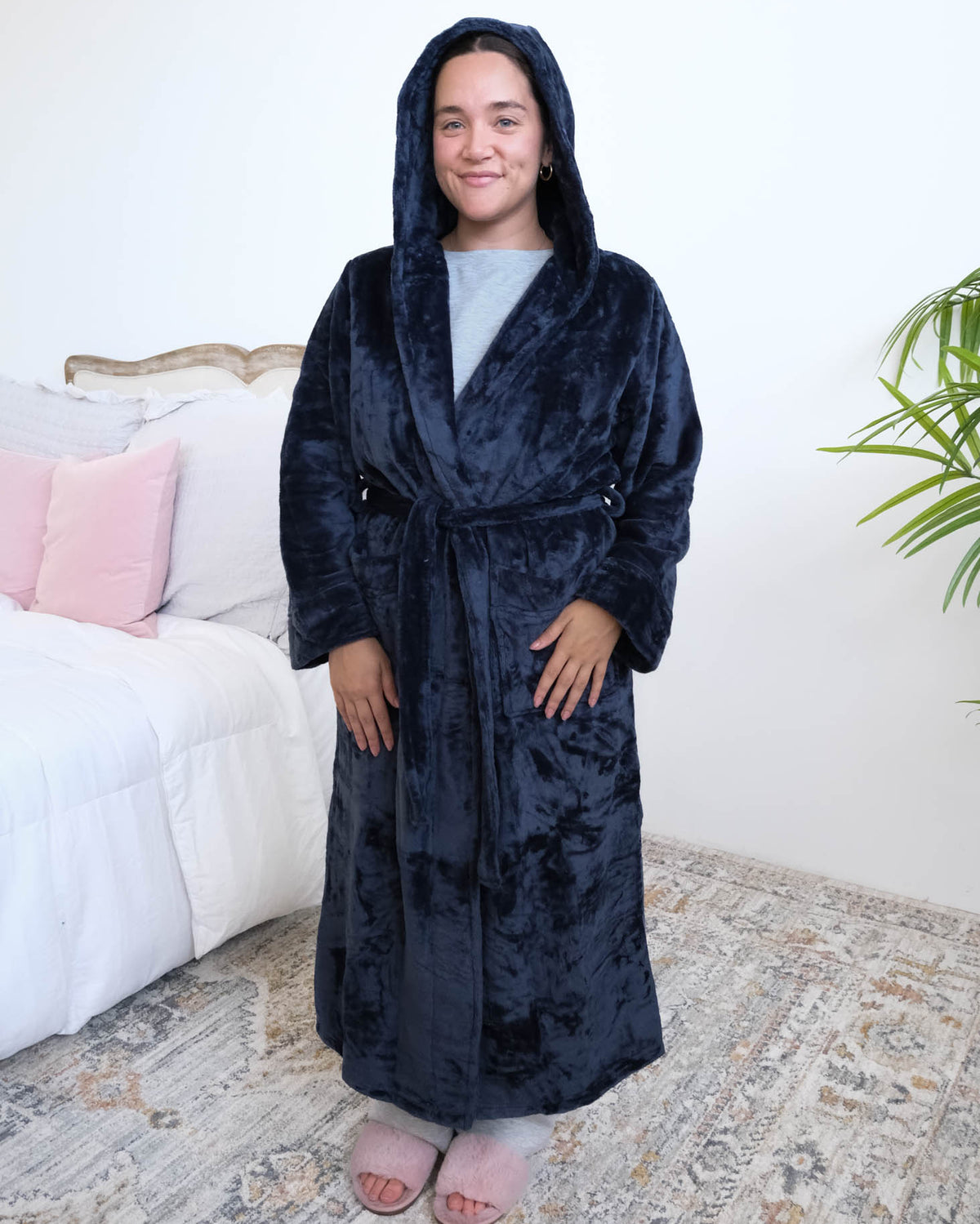 A woman wearing a plush navy blue hooded robe by Lounge By Jan Marie with the hood up on her head stands in a bedroom setting with a bed, rug, and plant visible in the background. Cloud Nine Pajamas Edmonton