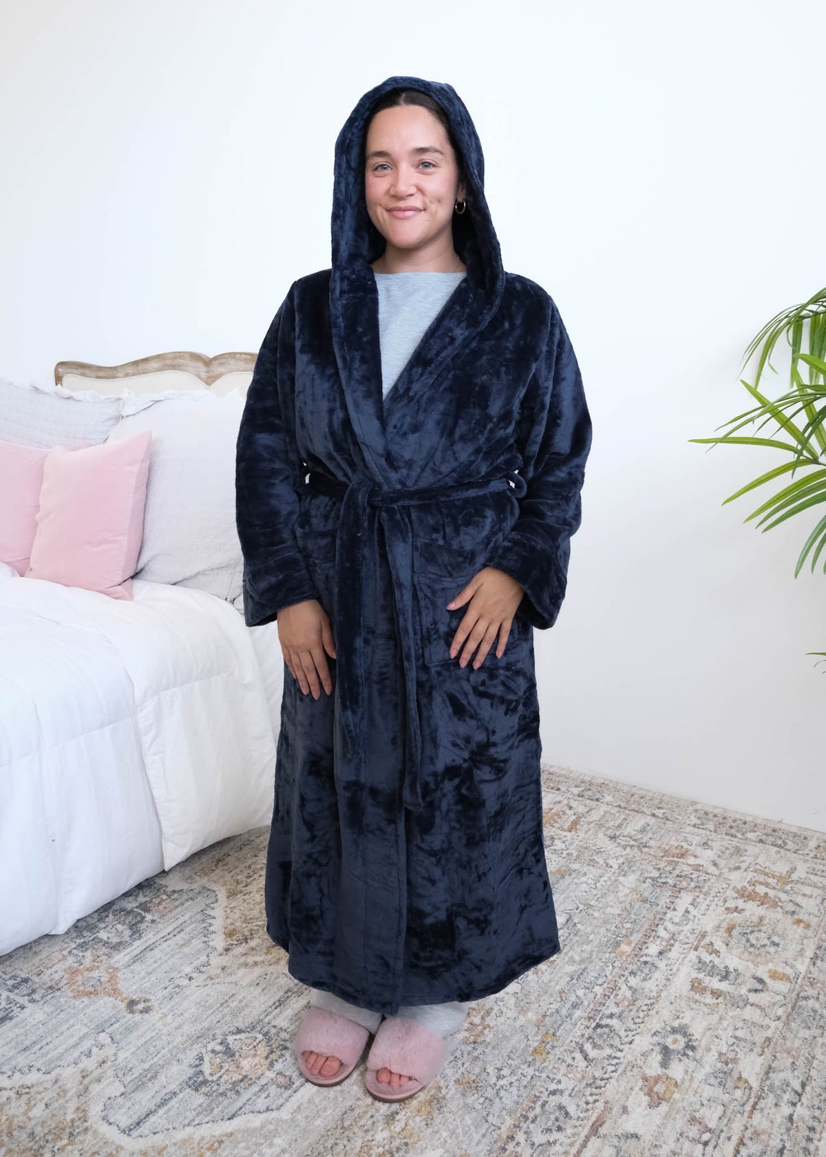 A woman wearing a plush navy blue hooded robe by Lounge By Jan Marie with the hood up on her head stands in a bedroom setting with a bed, rug, and plant visible in the background. Cloud Nine Pajamas Edmonton