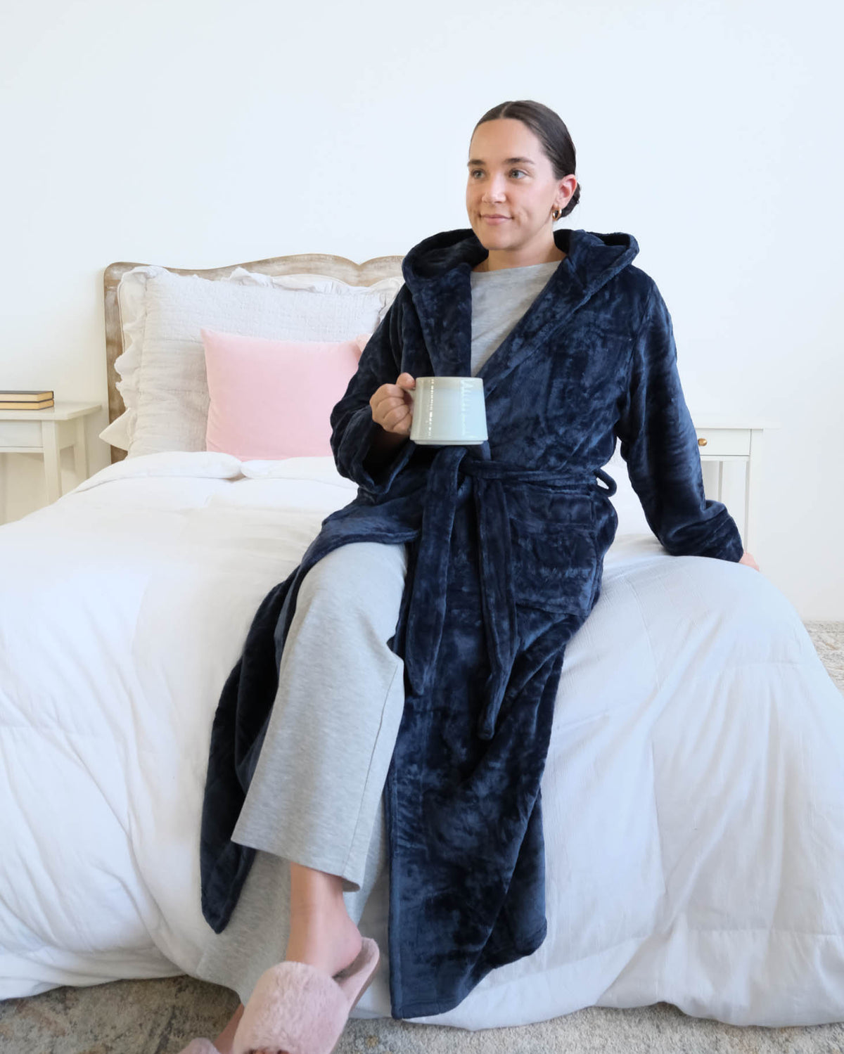 A woman sitting on a bed with a cup of coffee wearing a plush navy blue hooded robe by Lounge By Jan Marie in a bedroom setting with a bed. Cloud Nine Pajamas Edmonton