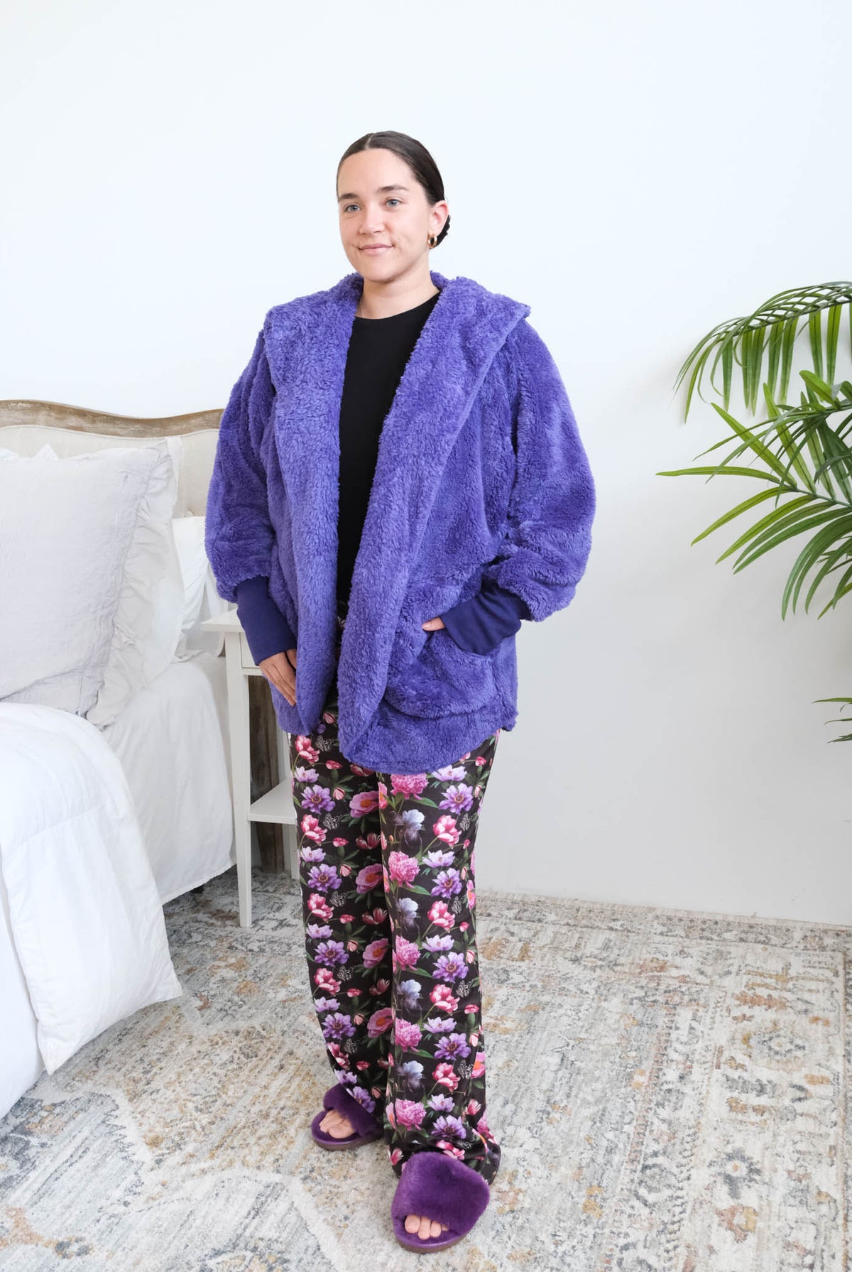 Woman standing in a bedroom wearing a blueberry purple fluffy hooded cardigan over a black top and floral pajama pants, with purple ribbed cuffs and purple fuzzy slippers, hands tucked into the cardigan's front pockets. Cloud Nine Pajamas Edmonton