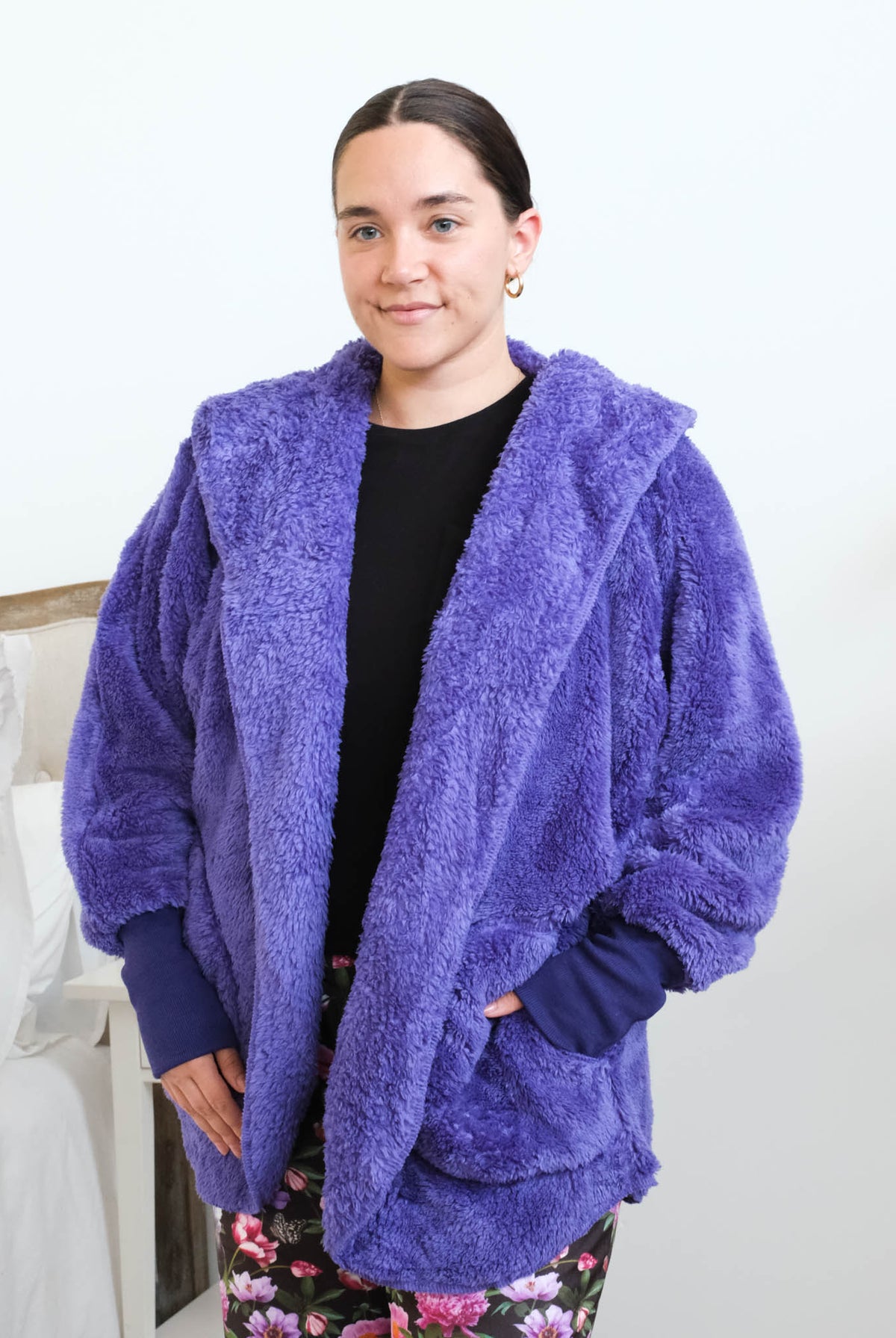 Close-up of a woman wearing the purple fluffy hooded by Lounge By Jan Marie cardigan open over a black top, showing the plush sherpa-like texture, wide lapel, and purple ribbed cuff at the wrist with one hand tucked into the front pocket. Cloud Nine Pajamas Edmonton