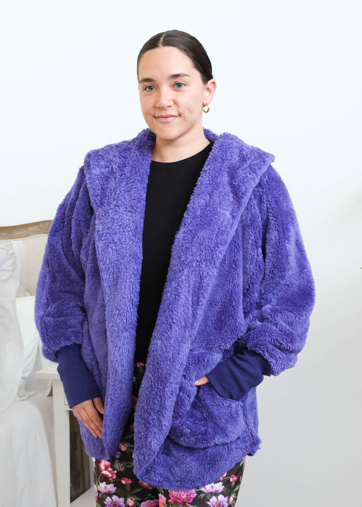 Close-up of a woman wearing the purple fluffy hooded by Lounge By Jan Marie cardigan open over a black top, showing the plush sherpa-like texture, wide lapel, and purple ribbed cuff at the wrist with one hand tucked into the front pocket. Cloud Nine Pajamas Edmonton