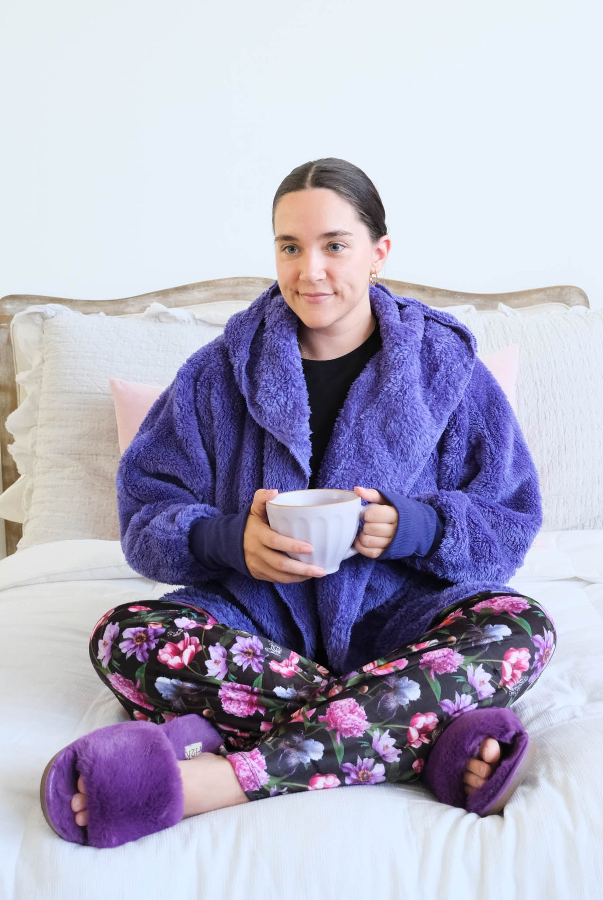 A woman sits cross-legged on a white bed wearing a flully plush sherpa purple hooded cardigan by Lounge by Jan Marie, floral pajama pants, and matching purple slippers while holding a mug. Cloud Nine Pajamas Edmonton