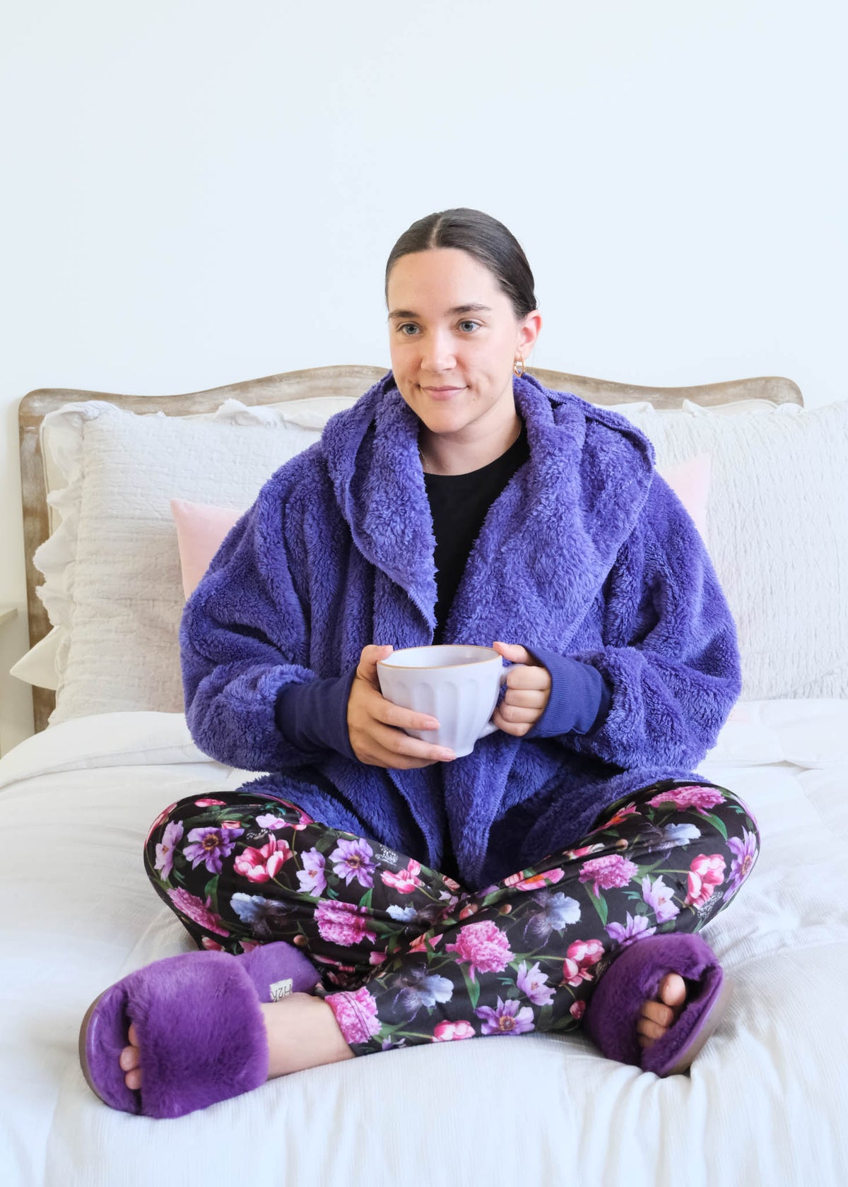 A woman sits cross-legged on a white bed wearing a flully plush sherpa purple hooded cardigan by Lounge by Jan Marie, floral pajama pants, and matching purple slippers while holding a mug. Cloud Nine Pajamas Edmonton
