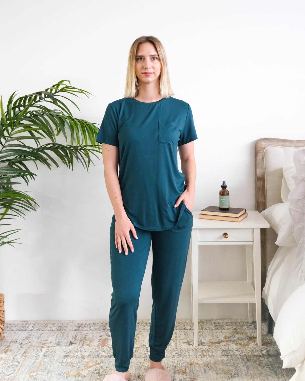 A woman modelling a matching two-piece bamboo dark teal loungewear set in a bright bedroom setting. The set consists of a short-sleeve crewneck top and matching jogger pants in a deep teal color. Cloud Nine Pajamas Edmonton