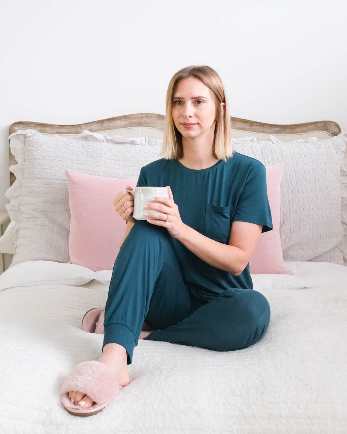 A woman sits on a bed wearing a dark teal short-sleeve pajama set with jogger style bottoms. She is wearing pink slippers, holding a white mug. The background features a white headboard, white bedding, and pink pillows. Cloud Nine Pajamas Edmonton