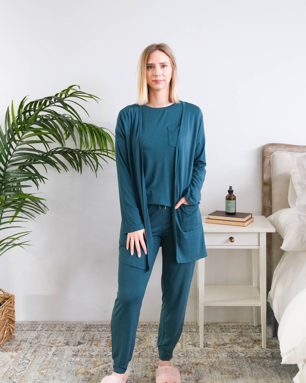 A woman modeling a matching bamboo dark teal lounge set consisting of a long-sleeve open-front cardigan, a long-sleeve top, and jogger pants, styled in a bright bedroom setting. Cloud Nine Pajamas Edmonton