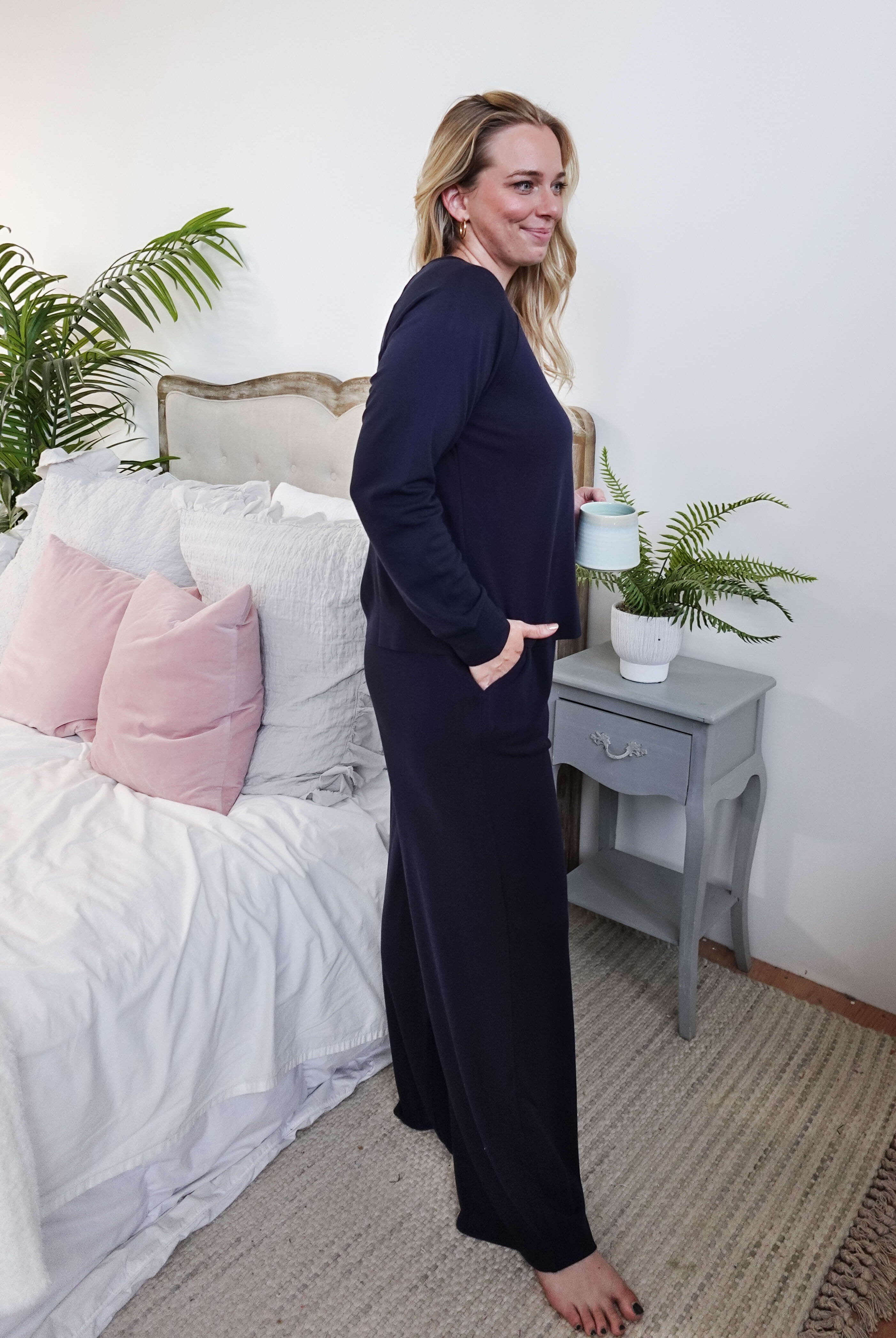Woman in Long Sleeve Raw Edge Straight Pant Lounge Set standing by a bed, holding a drink, with a relaxed smile.