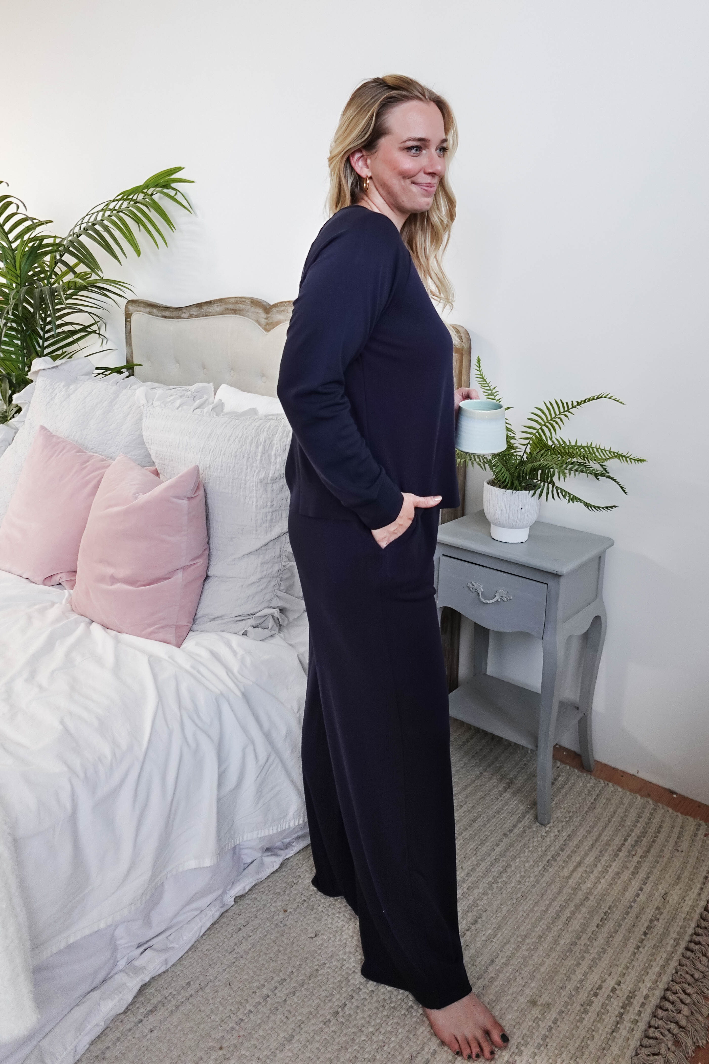 Woman in Long Sleeve Raw Edge Straight Pant Lounge Set standing by a bed, holding a drink, with a relaxed smile.