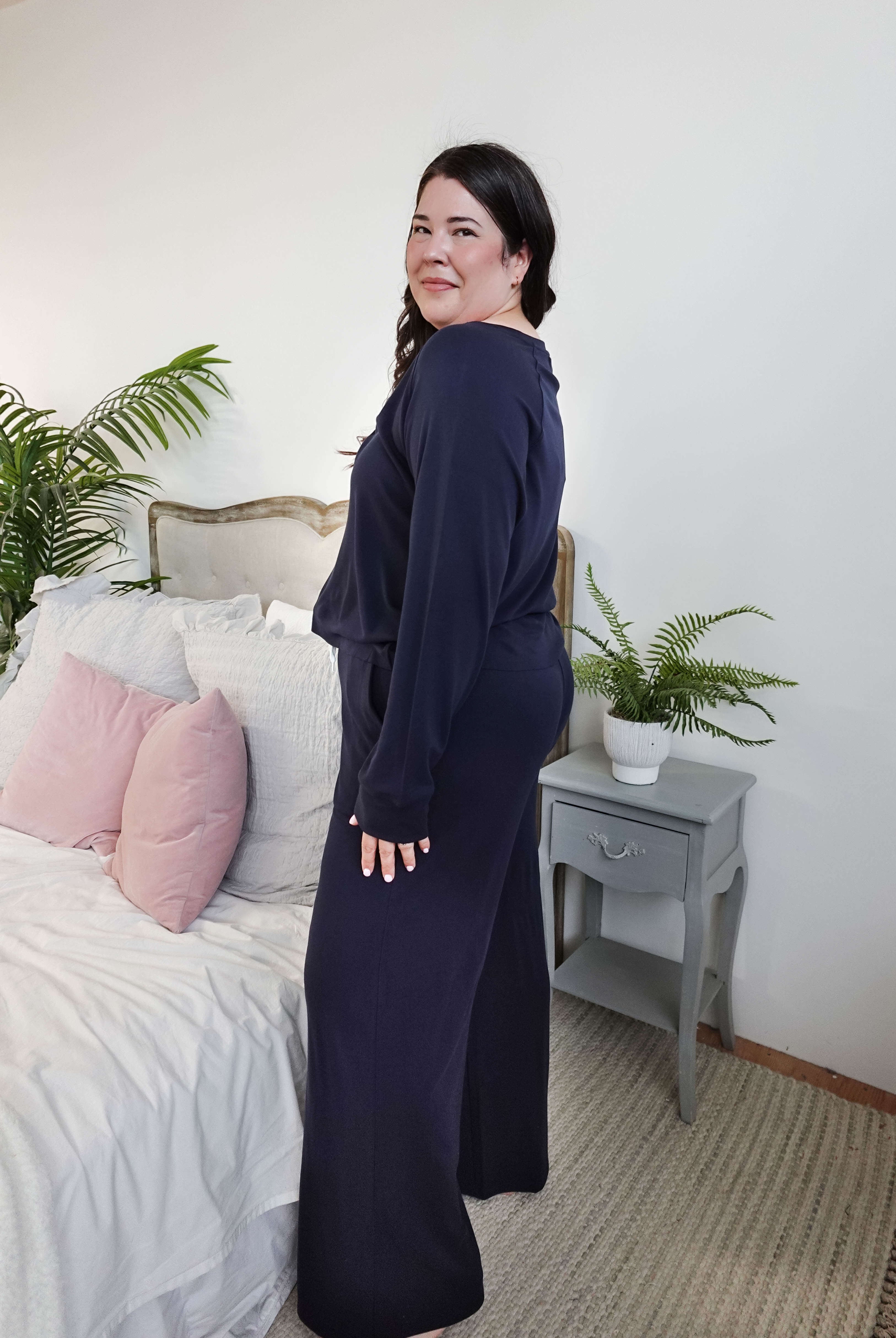 Model wearing Long Sleeve Raw Edge Straight Pant Lounge Set in navy, standing beside a bed with pink pillows and a plant.