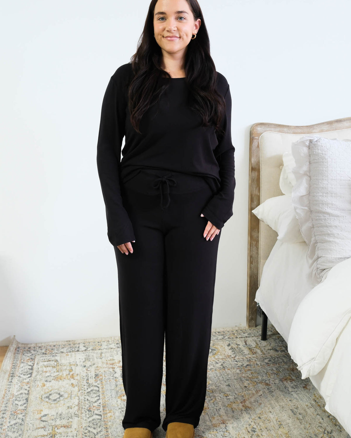 Model with long dark hair wearing a black long-sleeve scoop-neck pajama top half-tucked into matching black wide-leg pants with a drawstring tie waist, standing in a bedroom beside a white bed, styled with tan platform slippers. Cloud Nine Pajamas Edmonton.