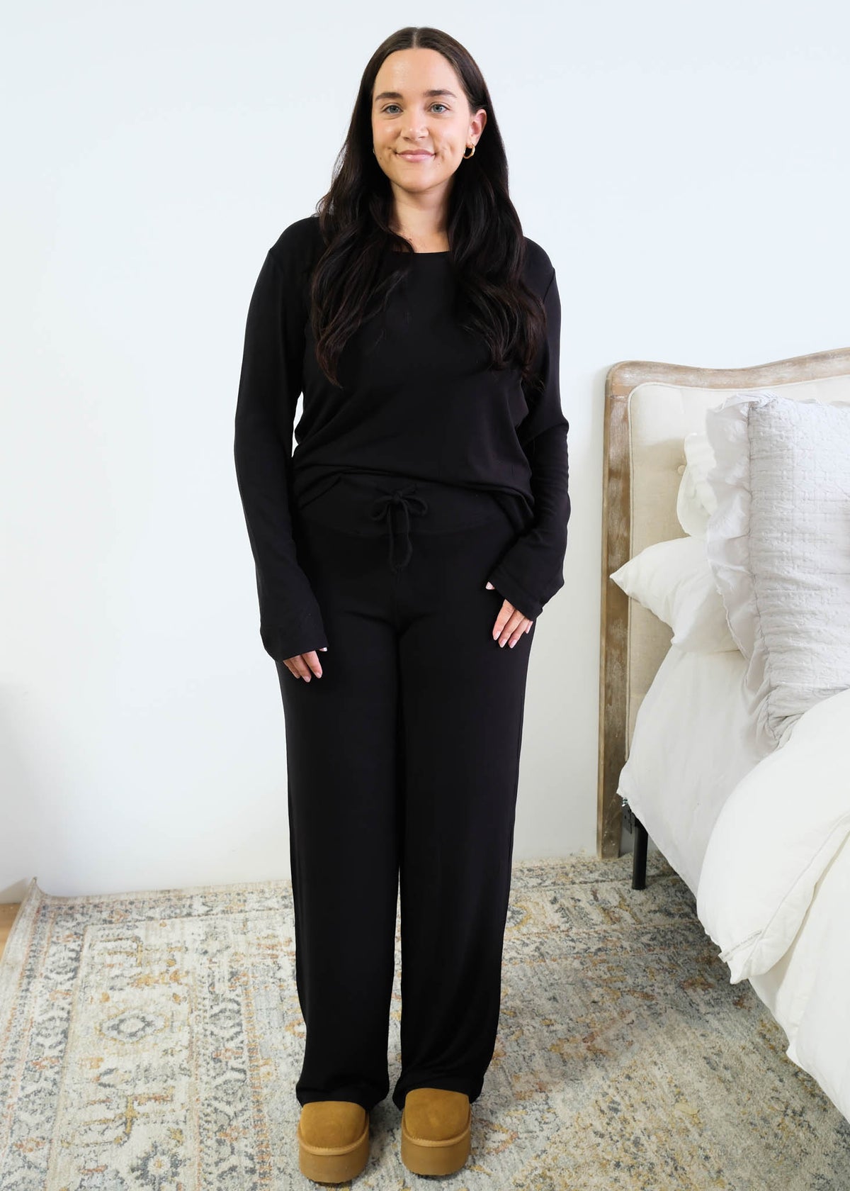 Model with long dark hair wearing a black long-sleeve scoop-neck pajama top half-tucked into matching black wide-leg pants with a drawstring tie waist, standing in a bedroom beside a white bed, styled with tan platform slippers. Cloud Nine Pajamas Edmonton.