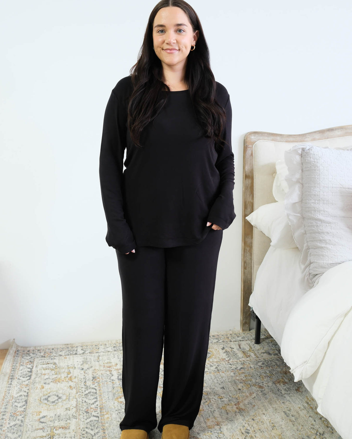 Front view of a black long-sleeve pajama top worn loose over matching black wide-leg lounge pants, showing the relaxed fit and curved hem, model standing beside a white bed with tan platform slippers. Cloud Nine Pajamas Edmonton.