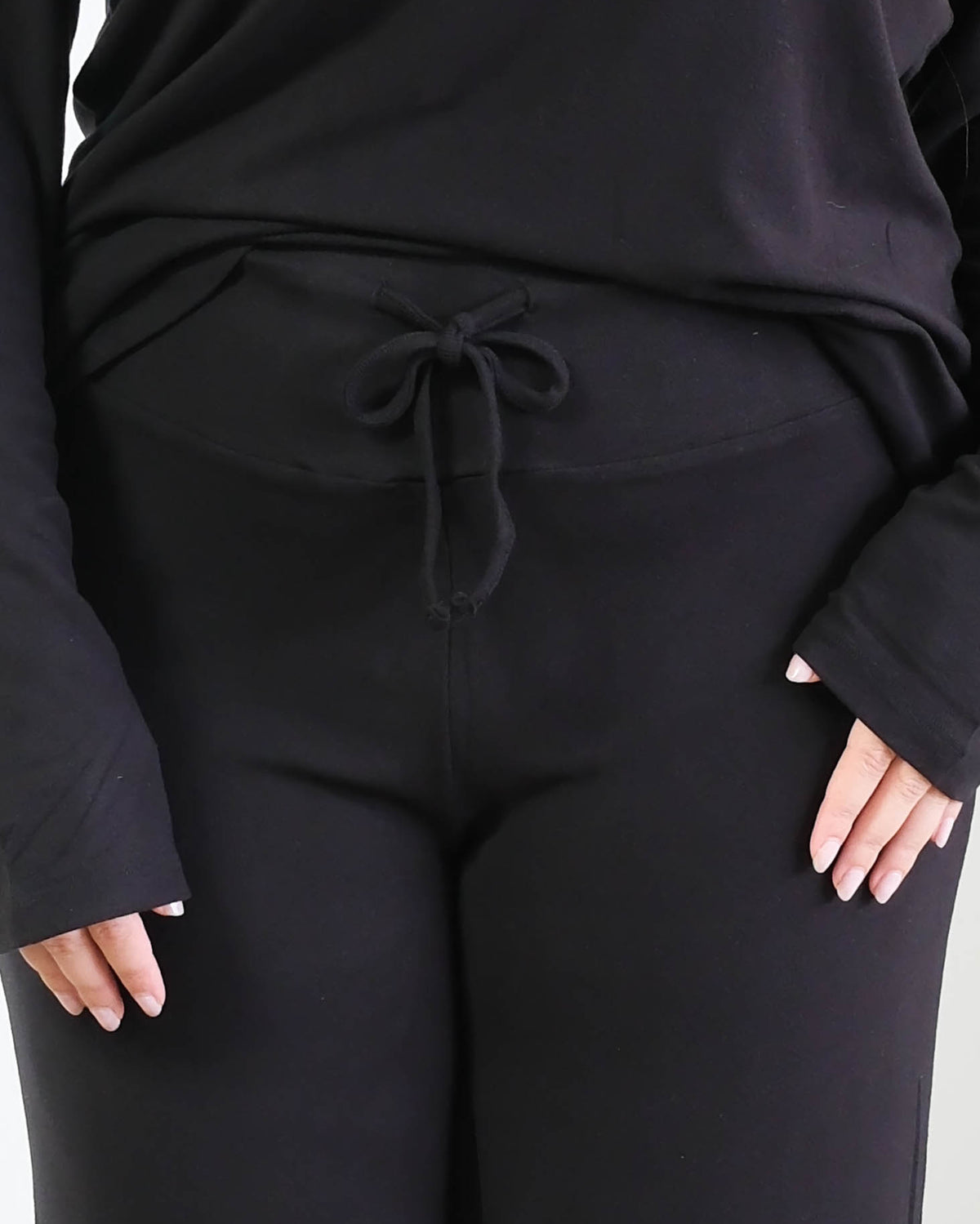 Close-up of black wide-leg pajama pants showing the wide, smooth fold-over waistband with a functional drawstring tied in a bow, worn with the matching black long-sleeve top. Cloud Nine Pajamas Edmonton.