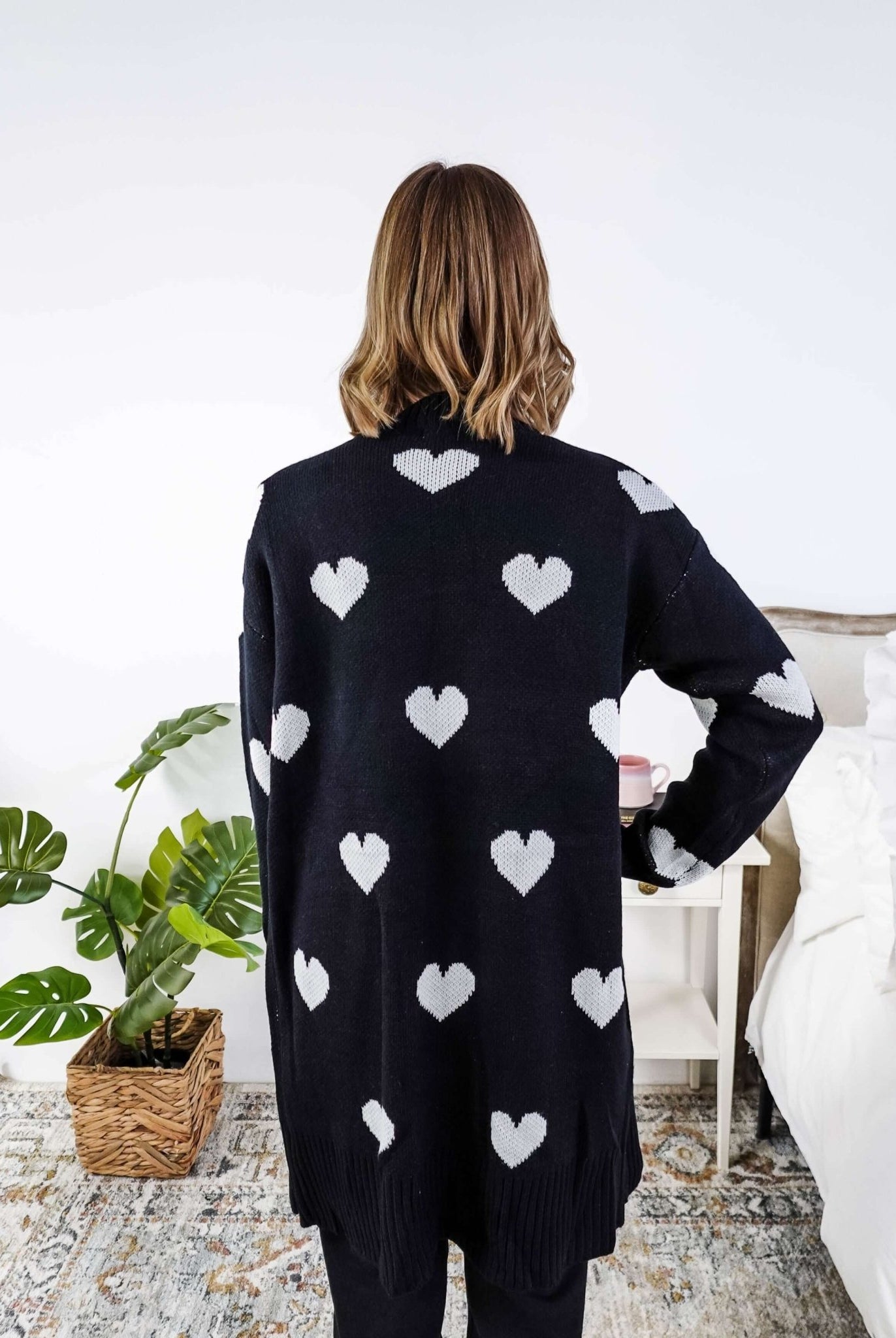 Women standing in a long black open cardigan with white hearts on it in a bedroom showing back of cardigan and the striking white hearts on a black background - Scarlett Ellie Black Hearts Knit Cardigan | Cloud Nine Pajamas