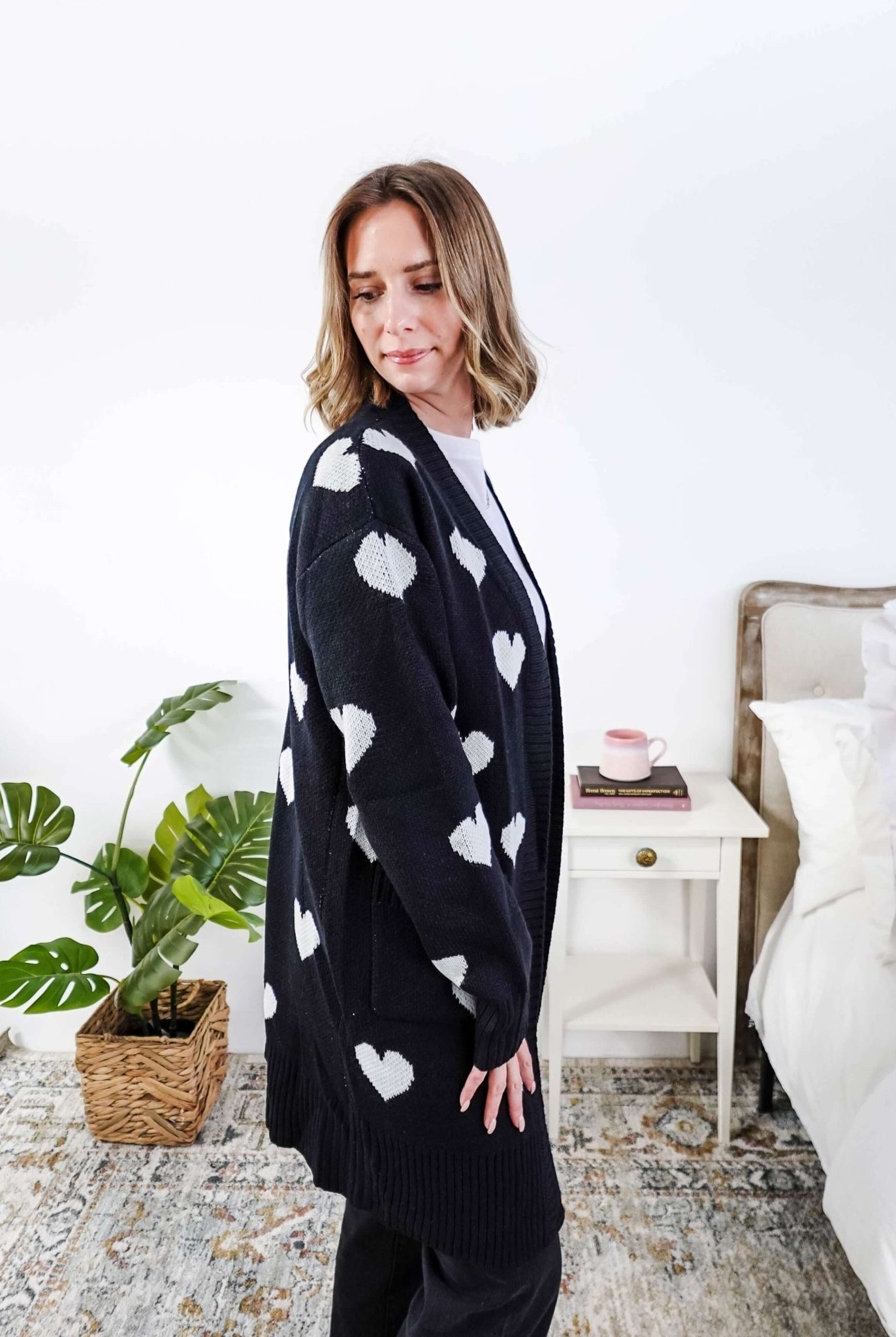 Scarlett Ellie Black Hearts Knit Cardigan | Cloud Nine Pajamas Women standing in a long black open cardigan with white hearts on it in a bedroom