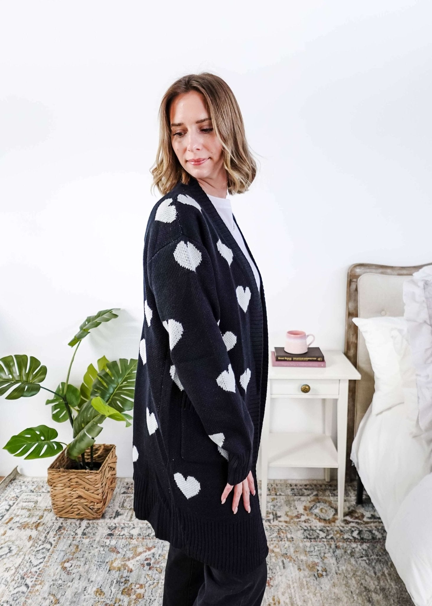 Scarlett Ellie Black Hearts Knit Cardigan | Cloud Nine Pajamas Women standing in a long black open cardigan with white hearts on it in a bedroom