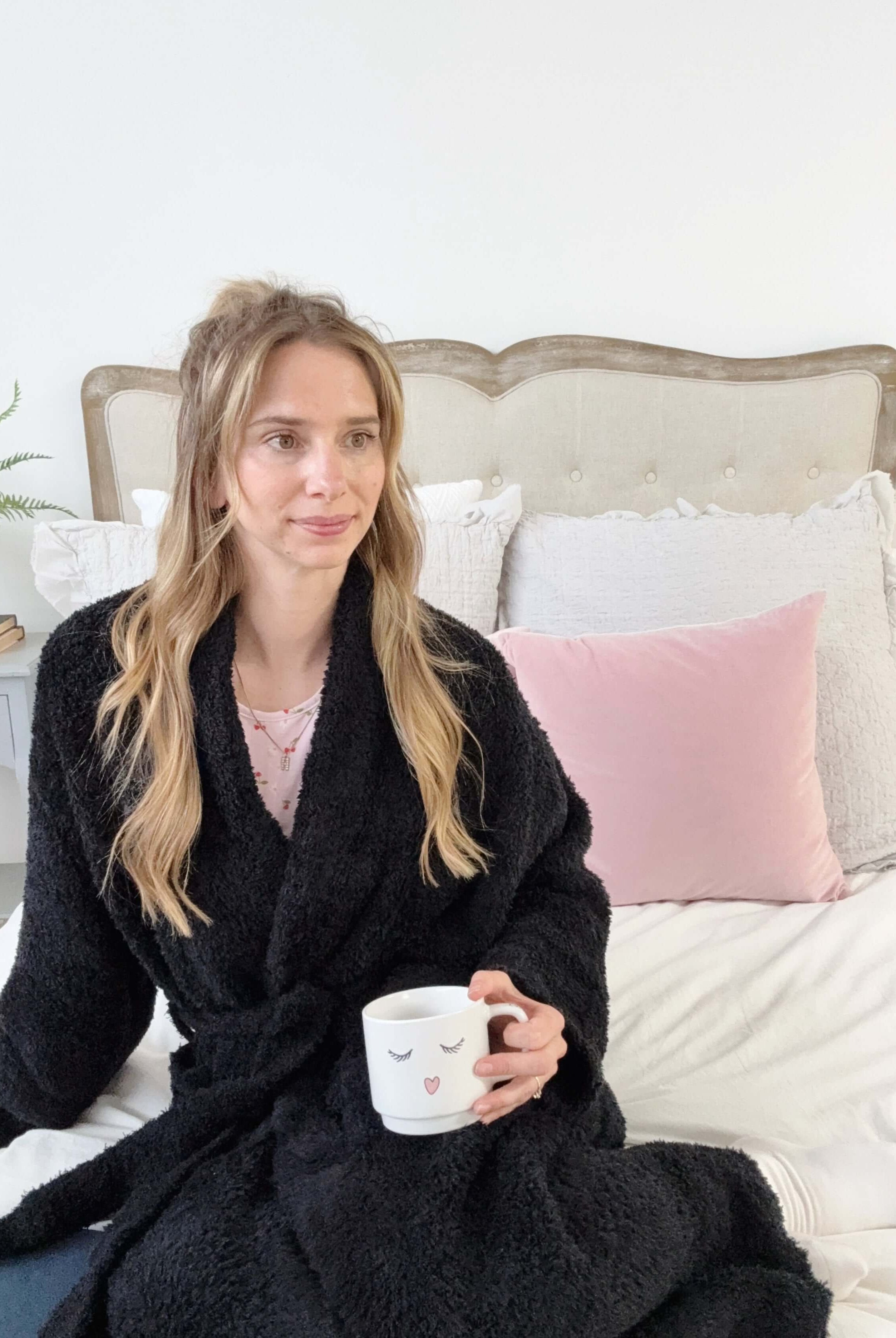 Woman in a black robe holding a mug on a bed with pink and gray pillows.