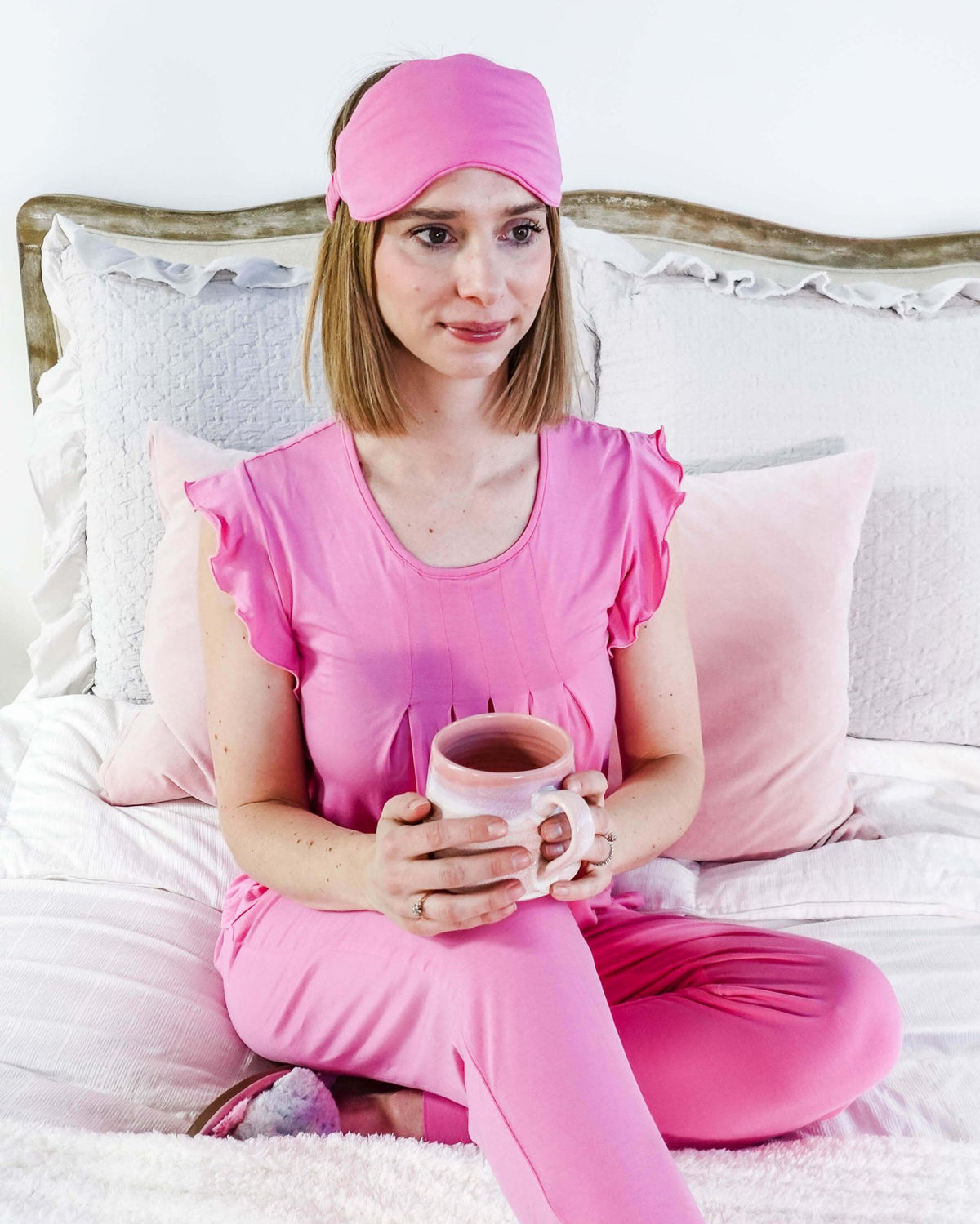 Woman wearing Bubblegum Pink Bamboo Sleep Eye Mask and pink pajamas, holding a mug while sitting on the bed.