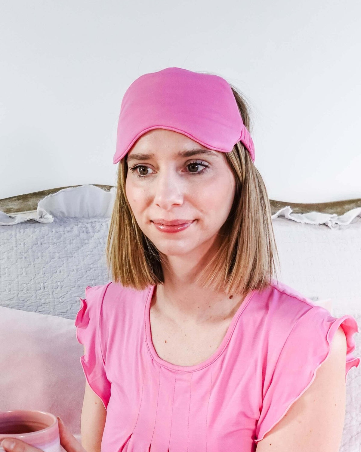 Woman wearing Bubblegum Pink Bamboo Sleep Eye Mask while holding a pink mug, sitting on a bed.