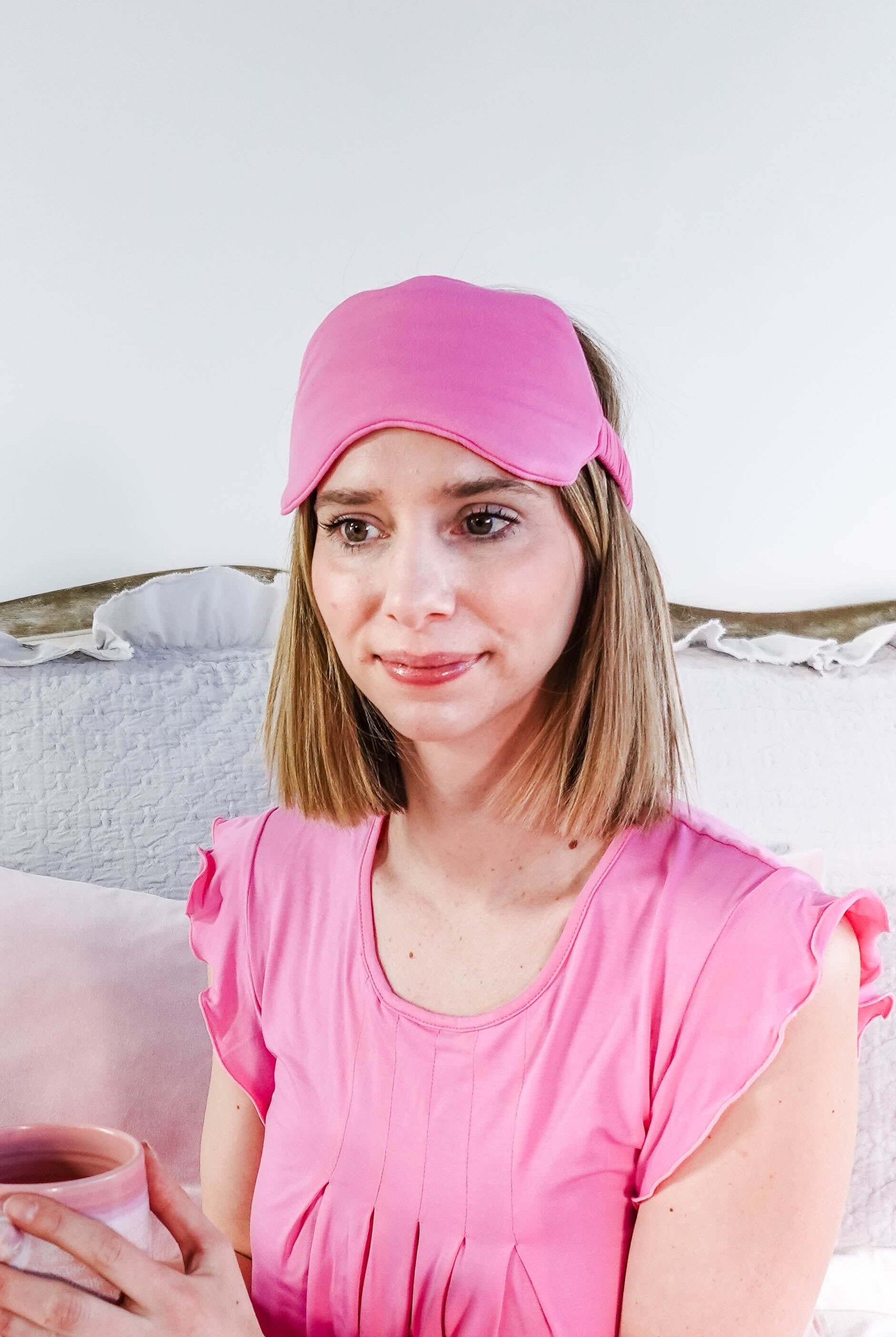 Woman wearing Bubblegum Pink Bamboo Sleep Eye Mask while holding a pink mug, sitting on a bed.