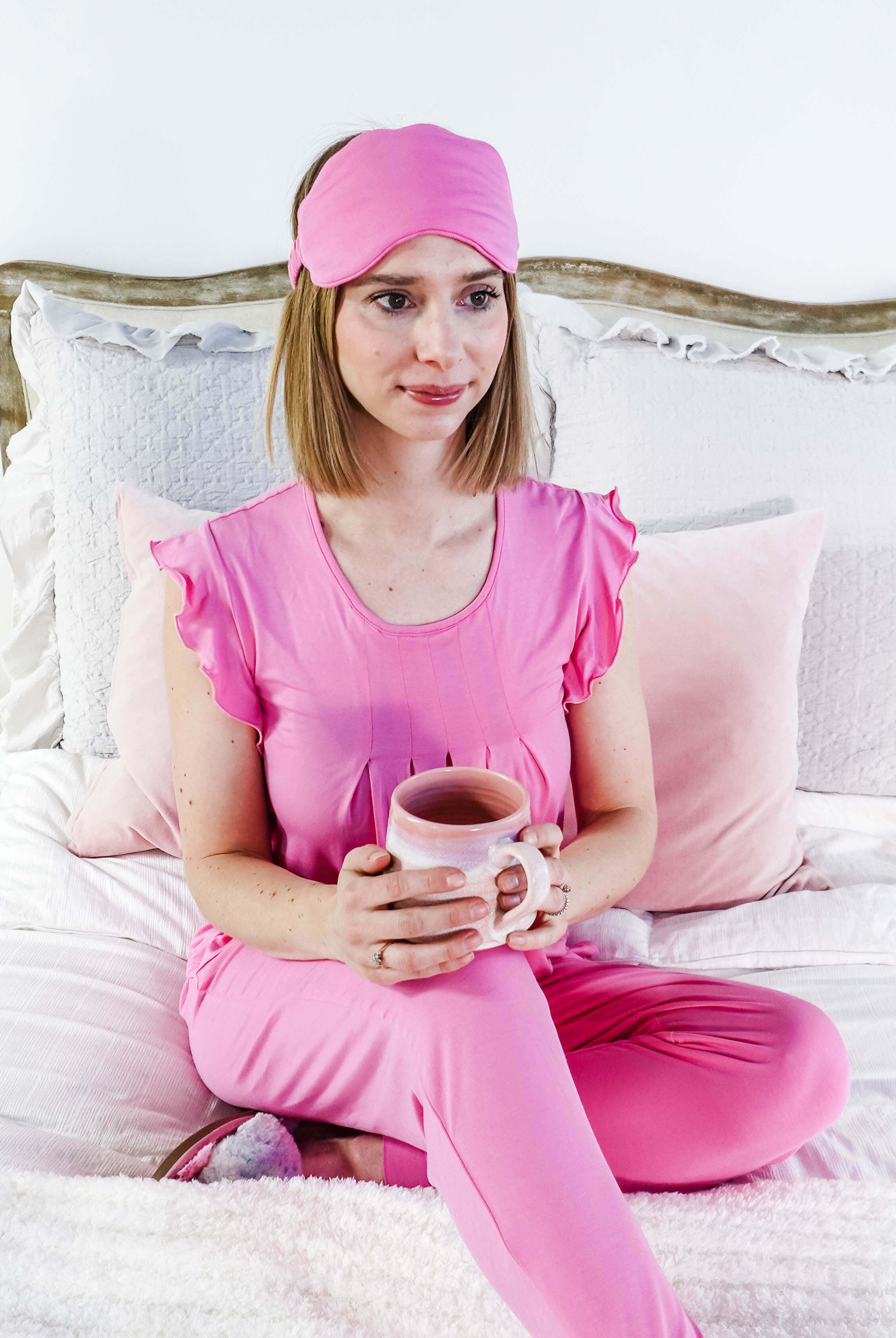 Woman wearing Bubblegum Pink Bamboo Sleep Eye Mask and pink pajamas, holding a mug while sitting on the bed.