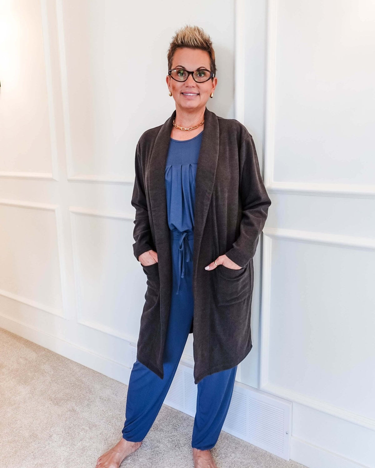 Full-length photo of a woman smiling, standing in a hallway, wearing a Charcoal Elevate Cardigan with deep patch pockets layered over a blue pyjama set. Classic collar, long-line structured fit.