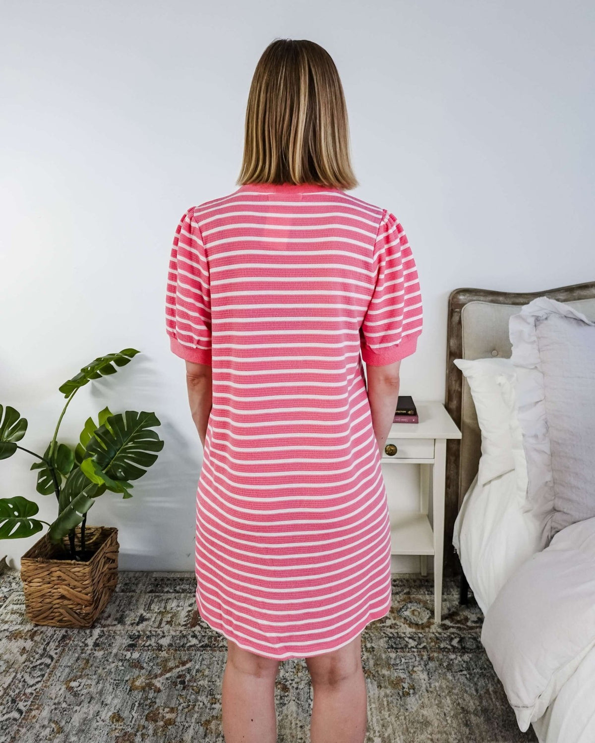 The back of a woman wearing a pink and white striped dress with puffed sleeves in a bedroom setting.