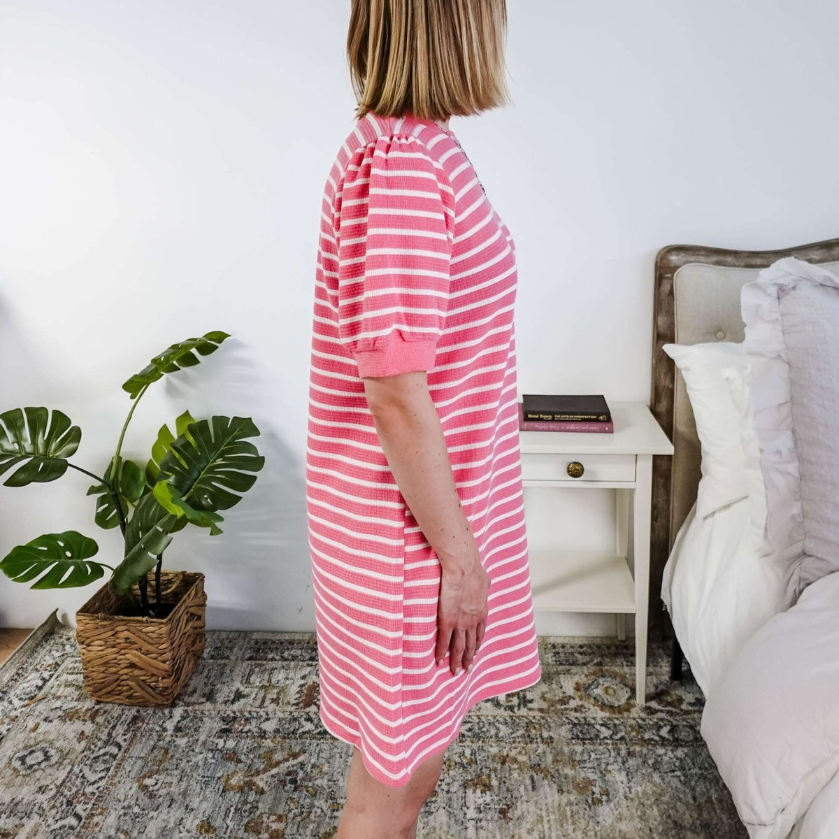The side of a woman wearing a pink and white striped dress with puffed sleeves in a bedroom setting.
