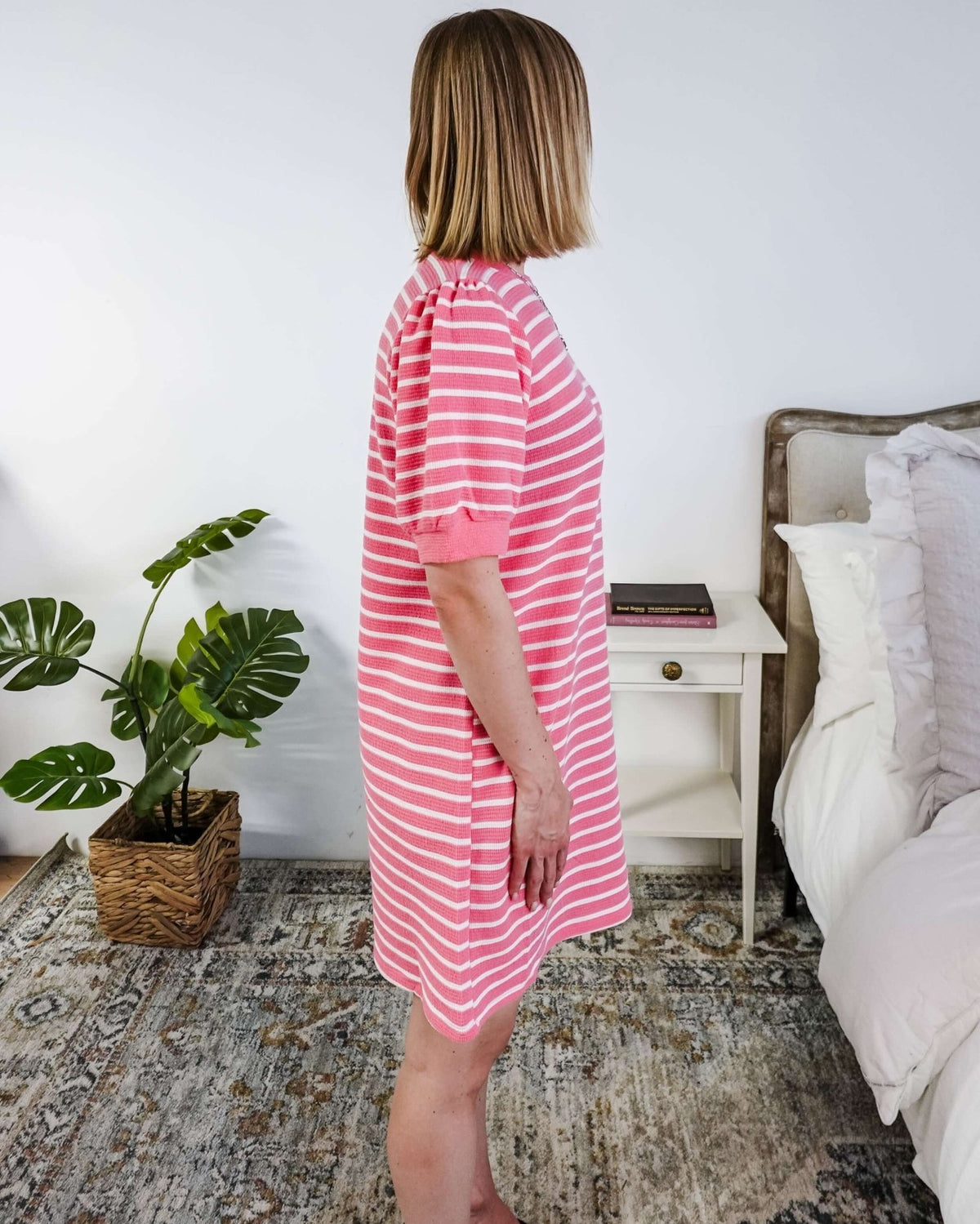 The side of a woman wearing a pink and white striped dress with puffed sleeves in a bedroom setting.