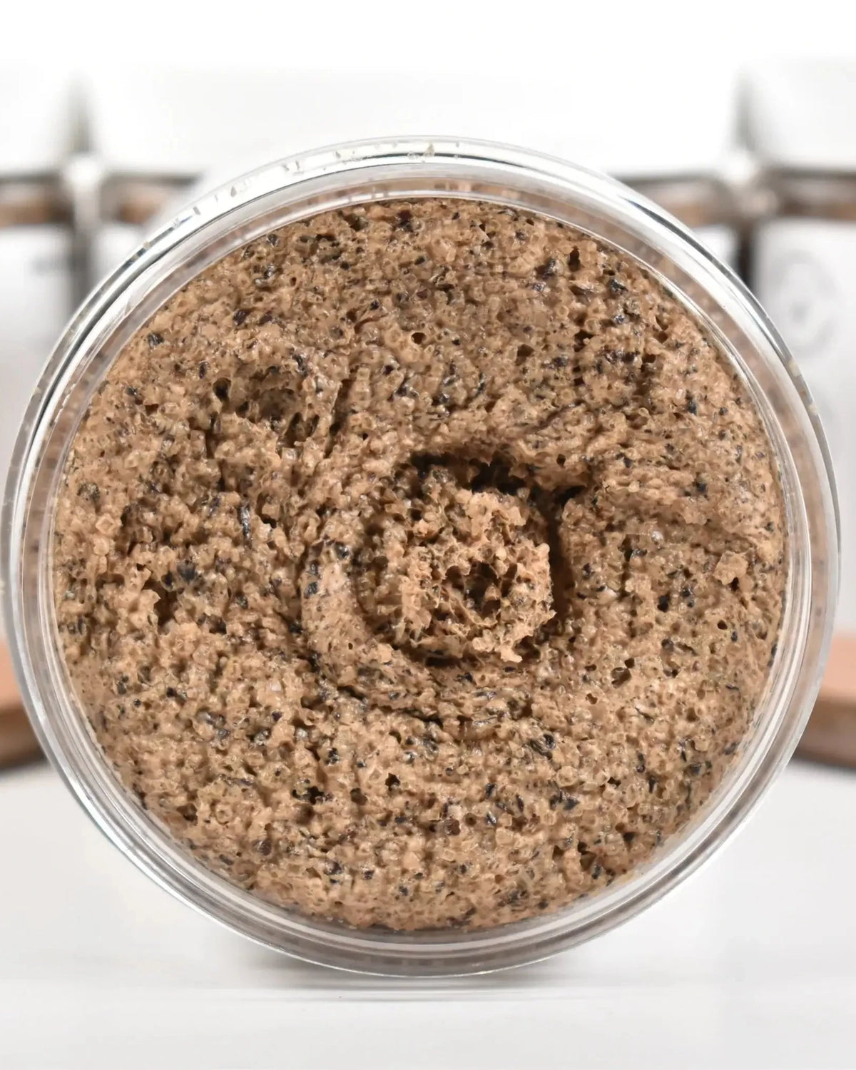 Coffee Bean Sugar Scrub in a clear jar showing the textured scrub, surrounded by blurred containers.