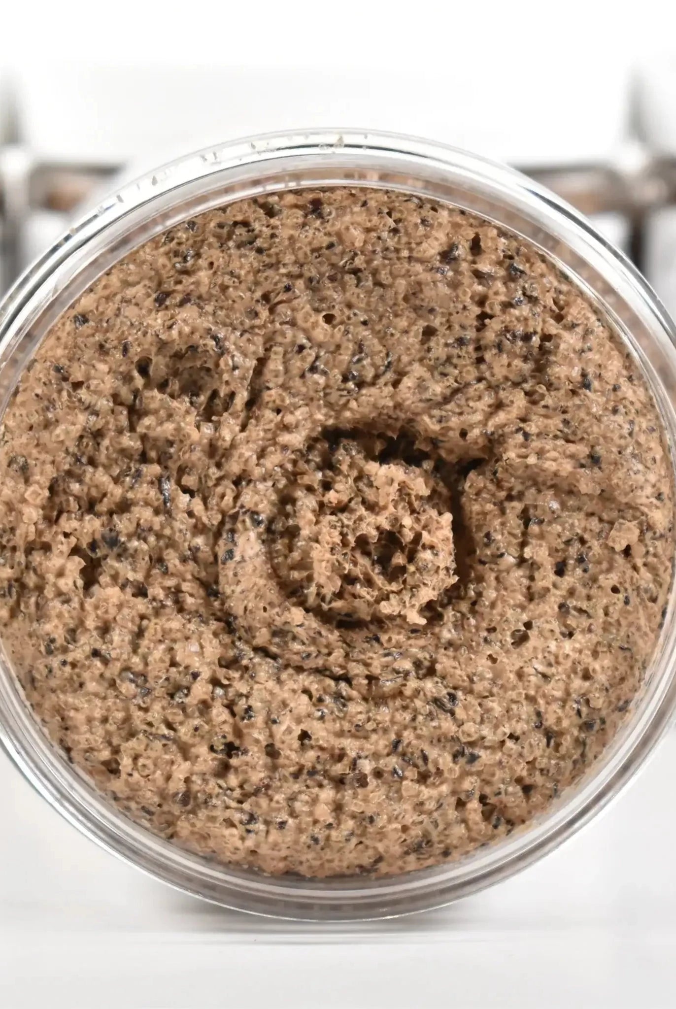 Coffee Bean Sugar Scrub in a clear jar showing the textured scrub, surrounded by blurred containers.