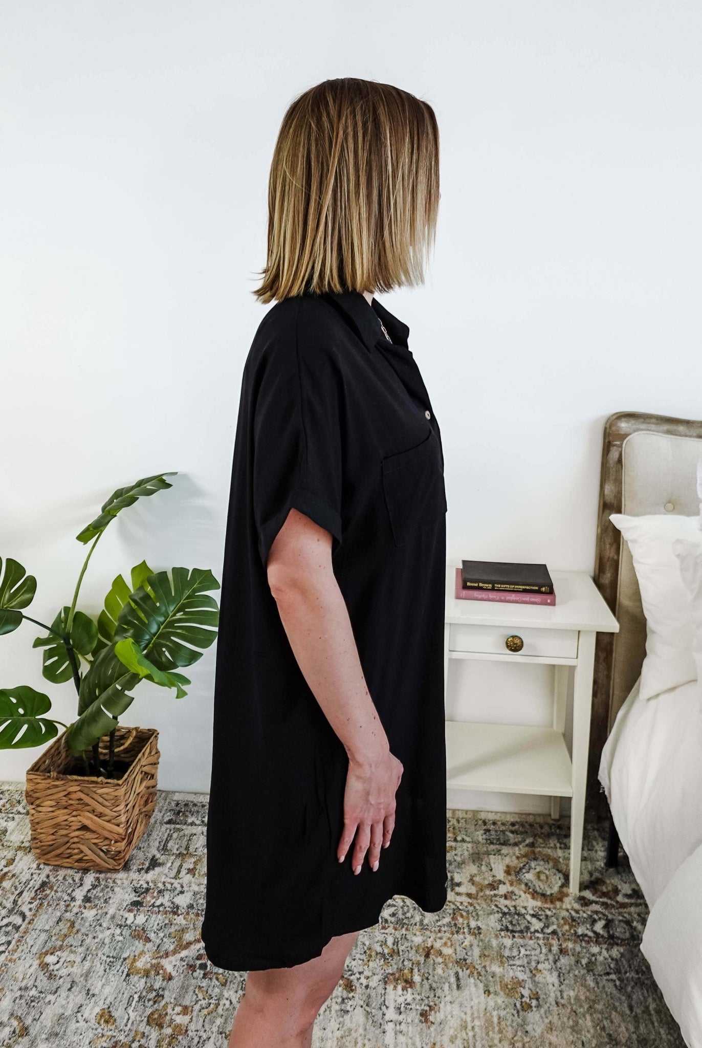 Woman wearing a black dress standing in a bedroom with a plant and nightstand. Scarlett Ellie Collared Button-Up Short Sleeve Dress | Cloud Nine Pajamas