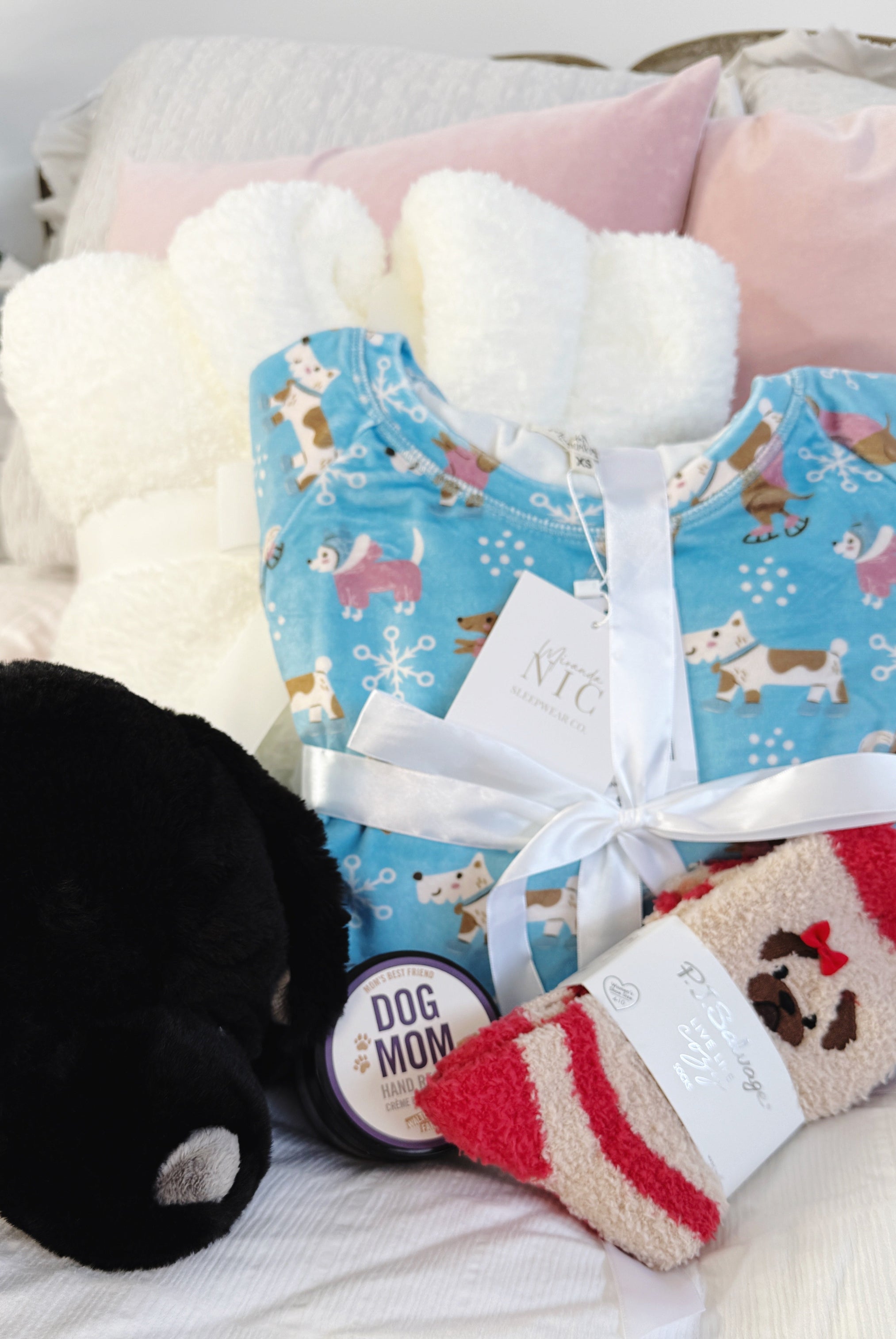 Dog Lover Gift Bundle Dog-themed gift set with a blue pj set with dogs skating print, red and beige pug socks, and a black plush labrador warming heat pack and hand rescue cream on a soft surface.
