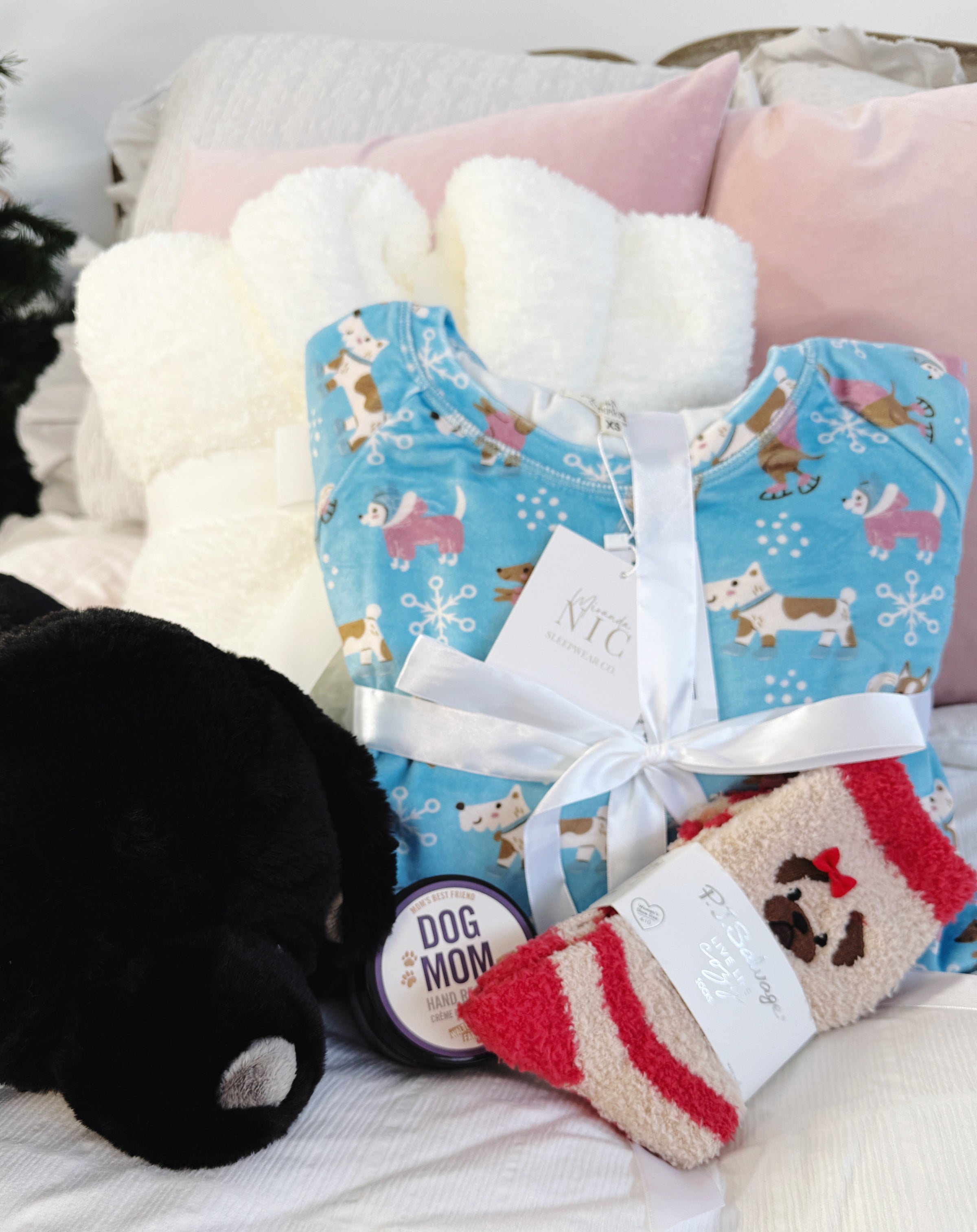Dog Lover Gift Bundle Dog-themed gift set with a blue pj set with dogs skating print, red and beige pug socks, and a black plush labrador warming heat pack and hand rescue cream on a soft surface.